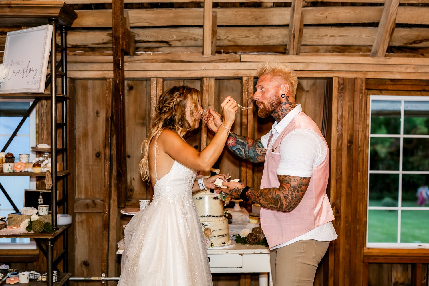 Bride and groom sharing cake cutting moment in rustic barn wedding venue, groom with tattoos wearing pink shirt.