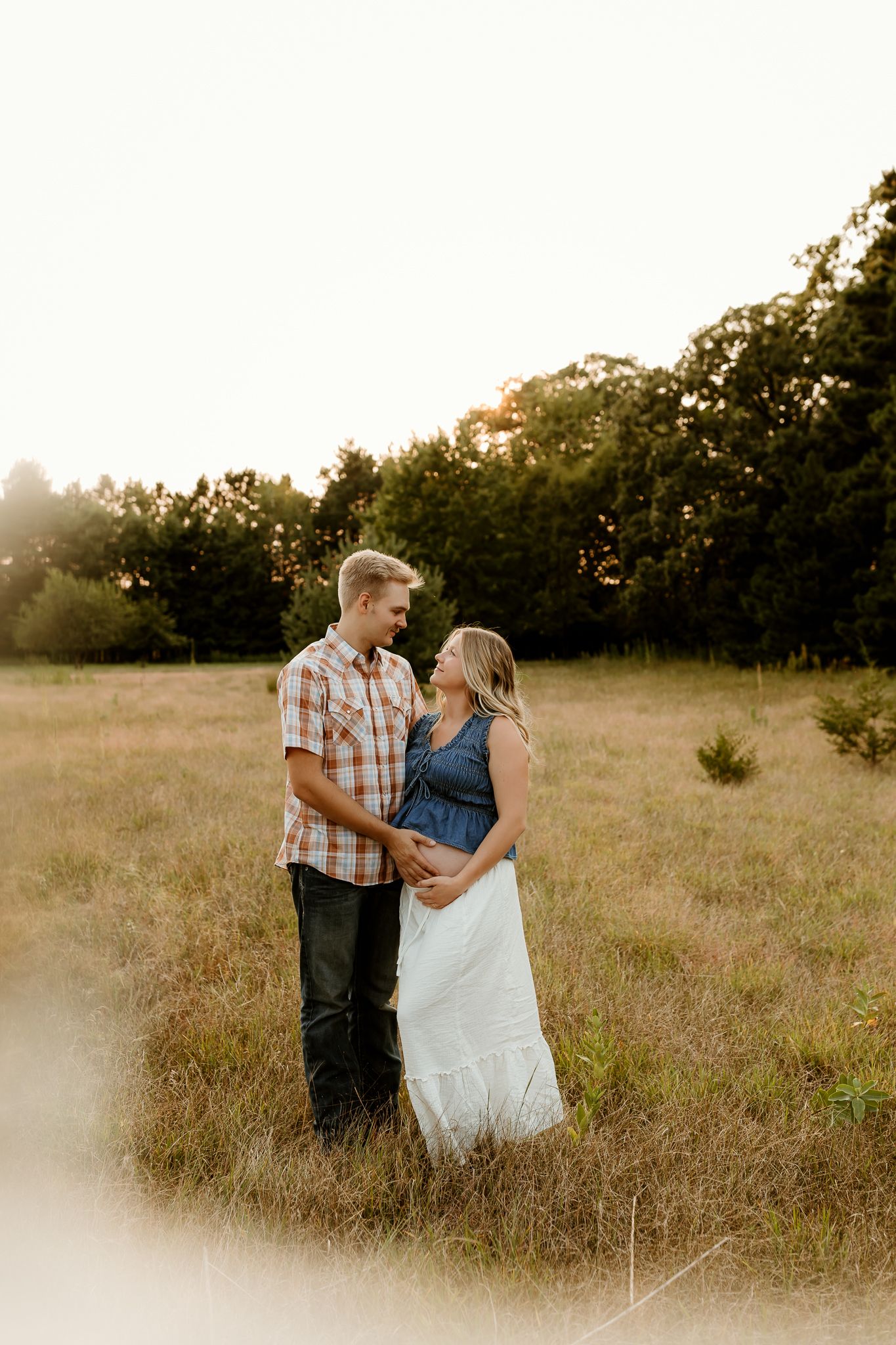 Couple shares a tender moment in a golden sunlit field, woman in white dress cradling baby bump.