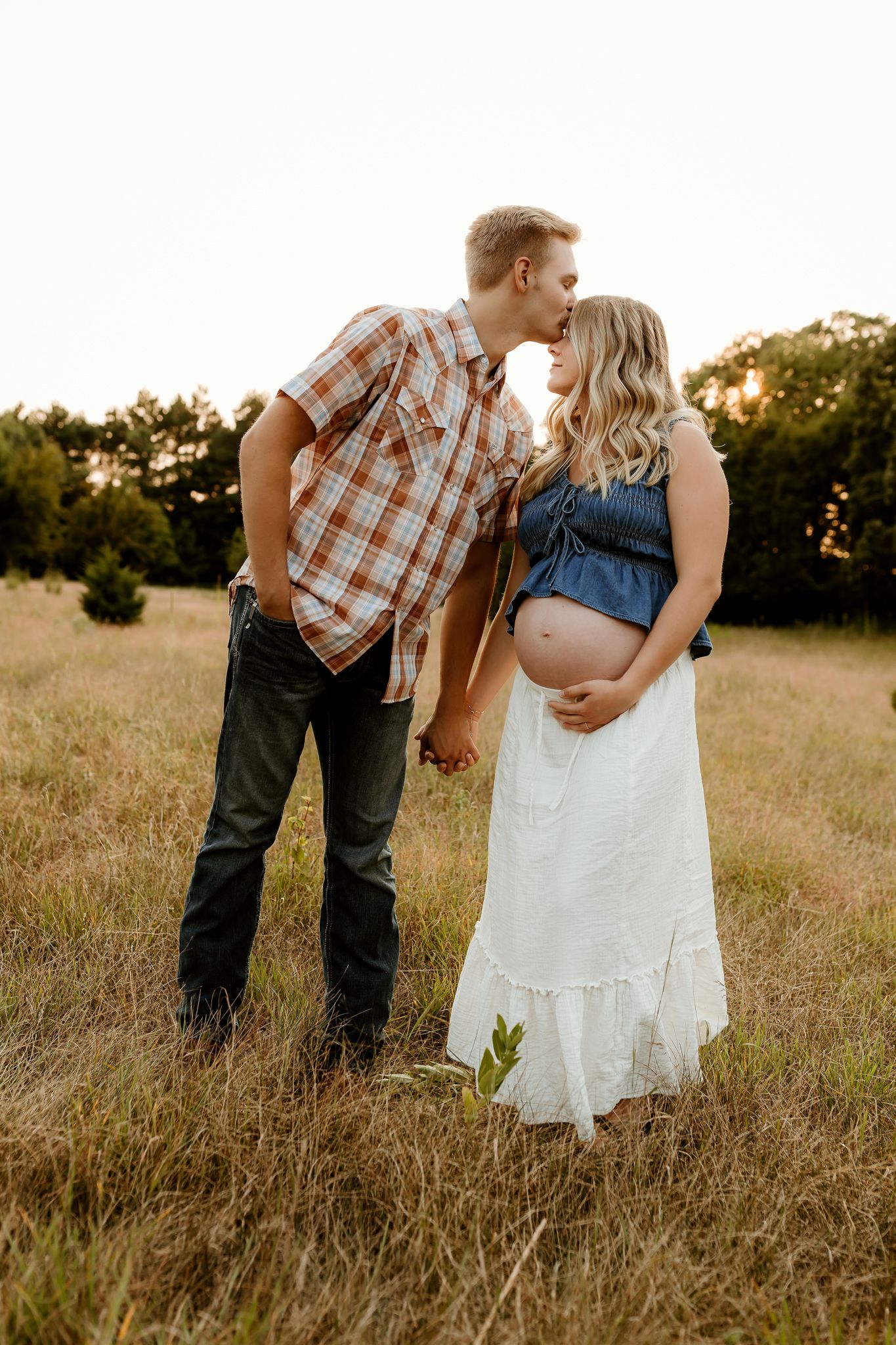 Couple sharing a kiss in a golden field, man in plaid shirt holds pregnant woman in denim top and white skirt.