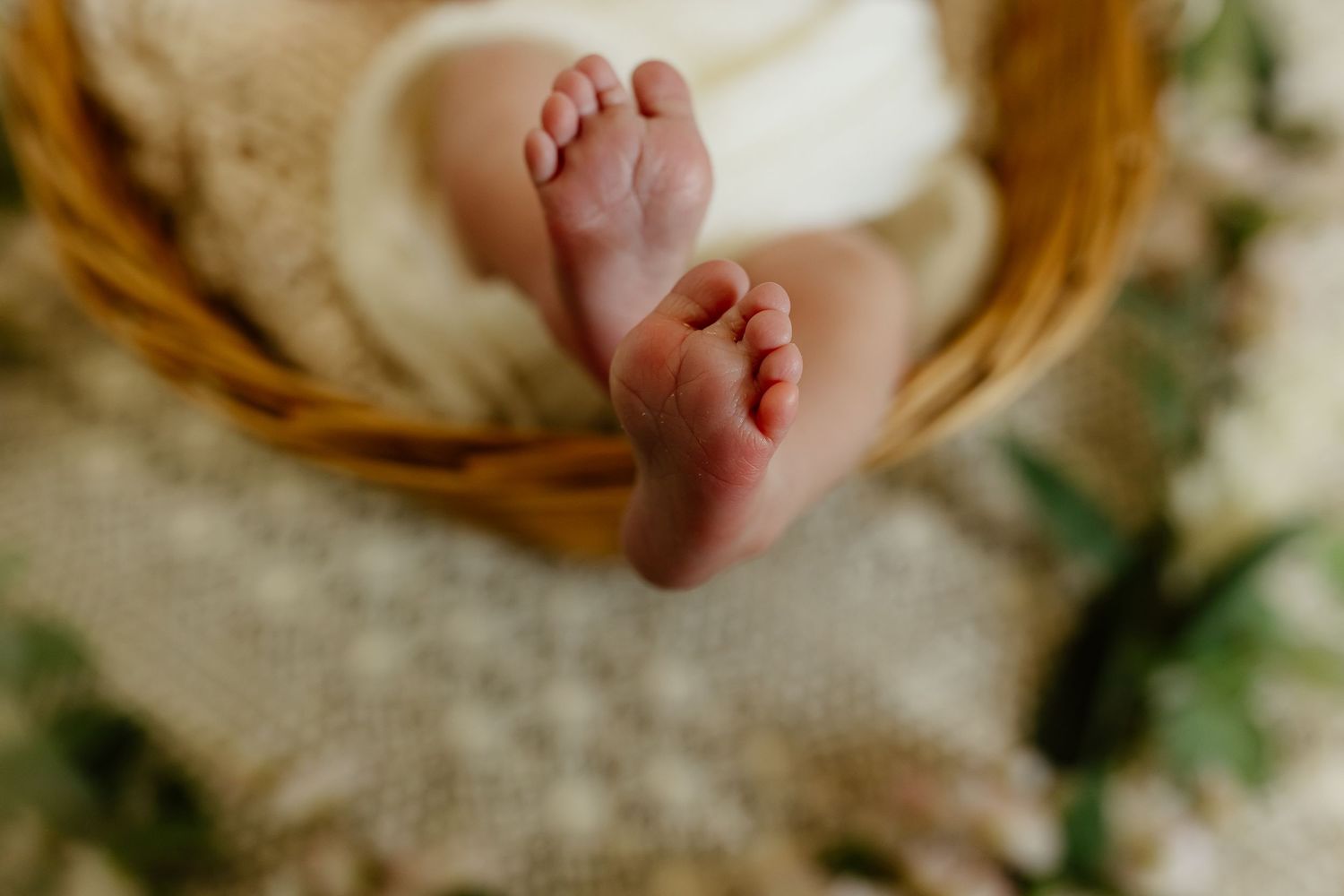 Close-up of newborn baby feet resting in a wicker basket with soft white blanket and flowers.