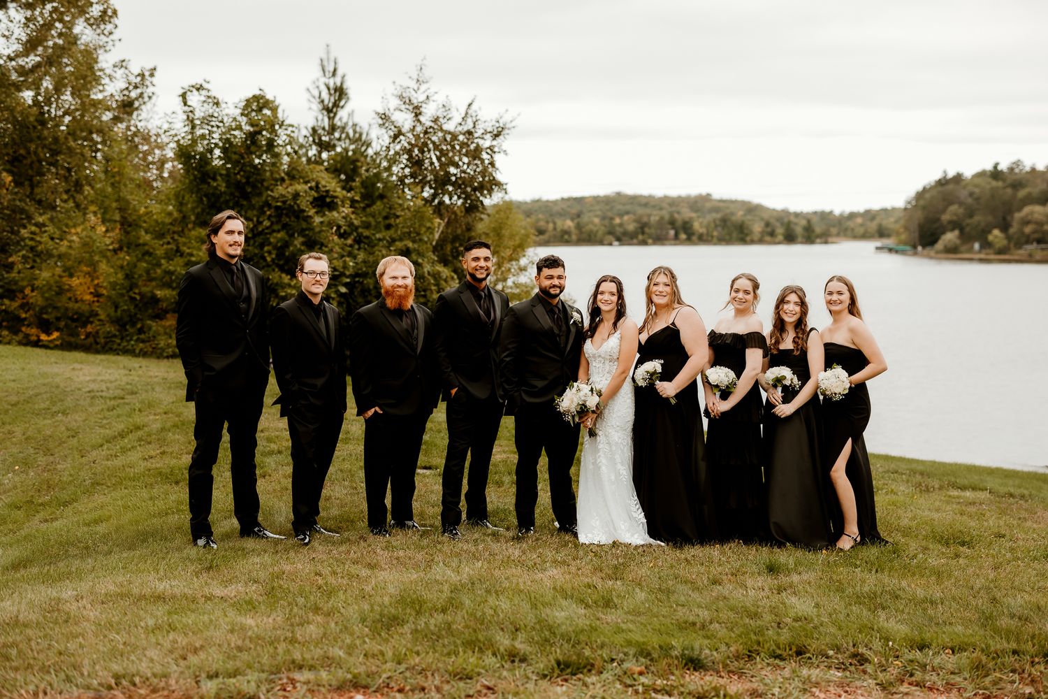Bride in white gown holding a bouquet poses with bridesmaids in black dresses and groomsmen in black suits on a grassy lakeside lawn.