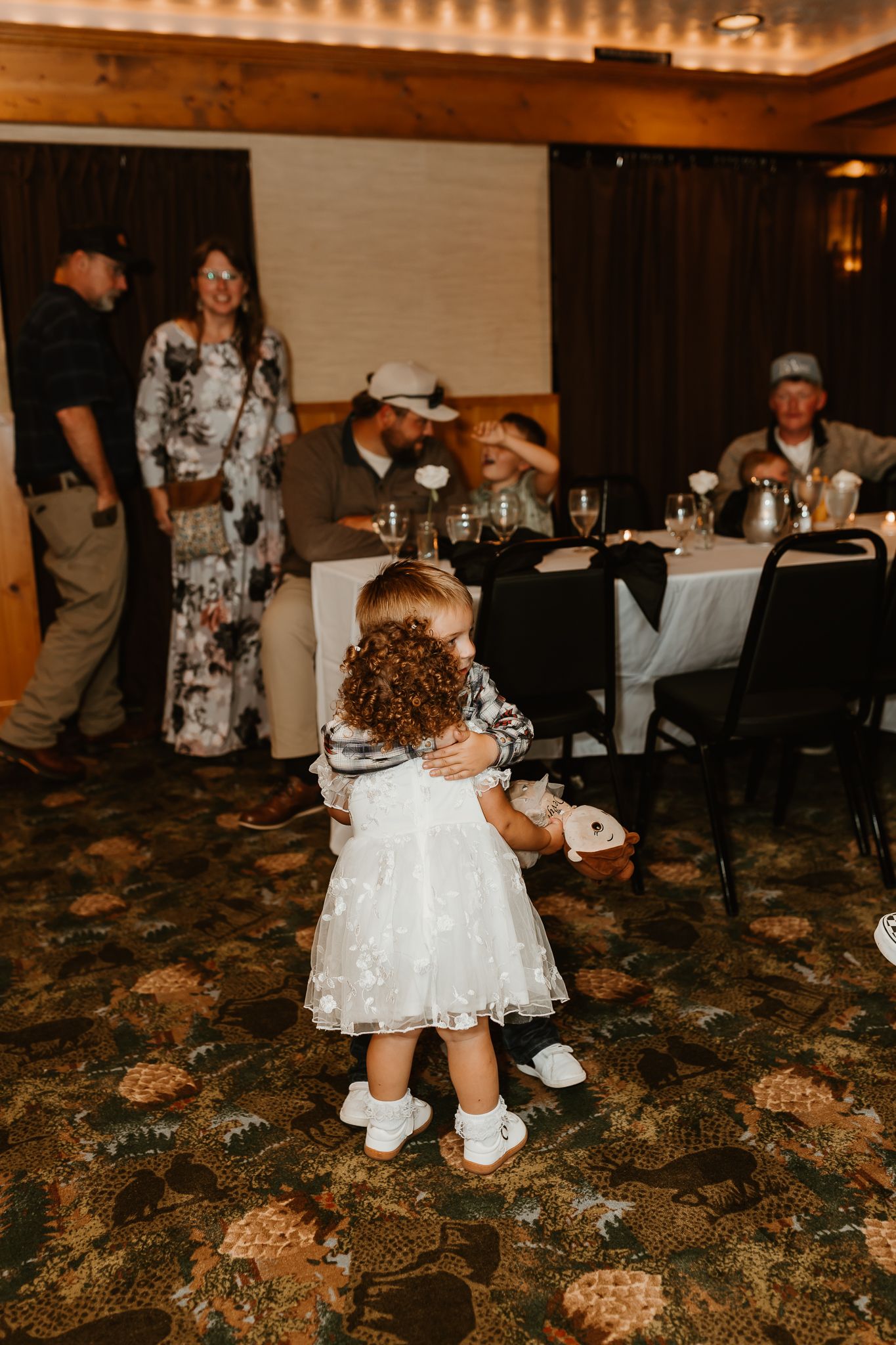 Two toddlers in white dresses hug on a patterned carpet at a banquet reception while guests sit and stand near tables in the background.
