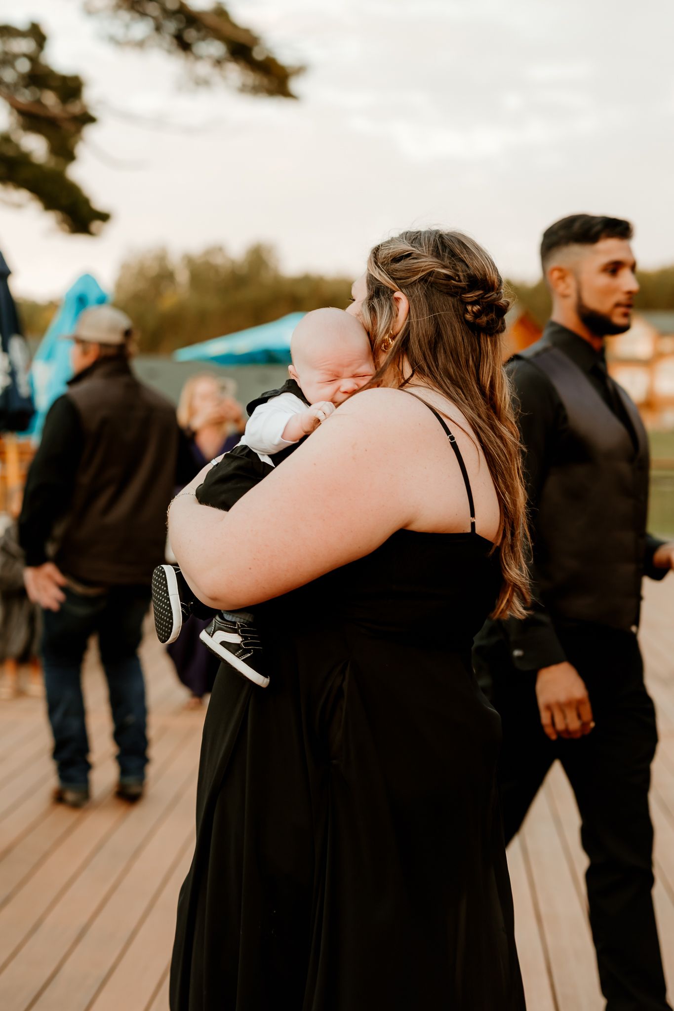 A woman in a black dress with a braided hairstyle holds a sleeping baby on her shoulder on a wooden deck at an outdoor event.