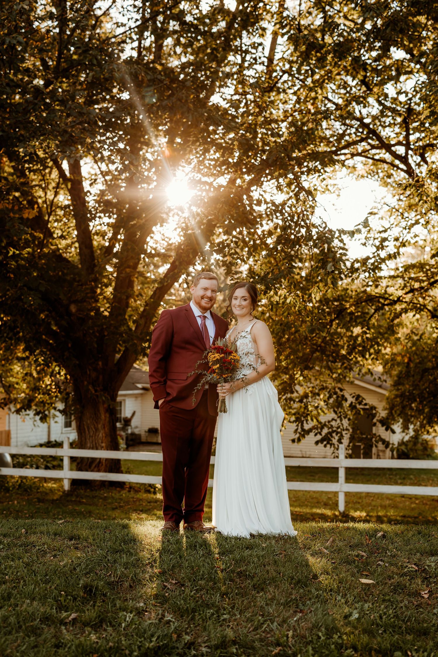 Smiling bride in a white gown and groom in a burgundy suit hold a bouquet under a large tree in golden sunset light.