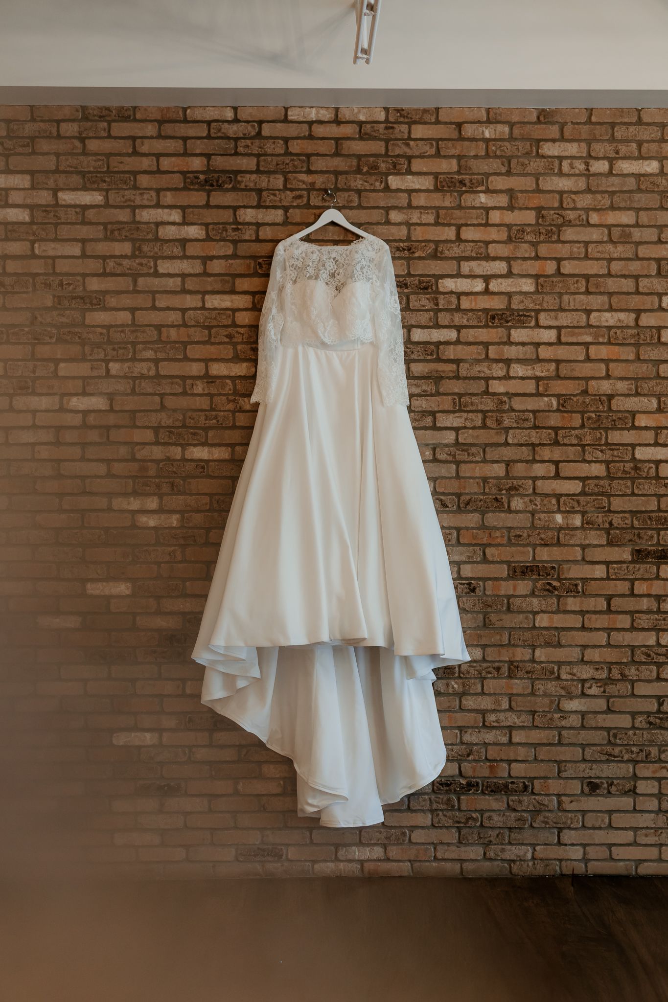 White lace long-sleeve wedding dress with a flowing satin skirt and high-low train hanging on a hanger against a rustic exposed brick wall.