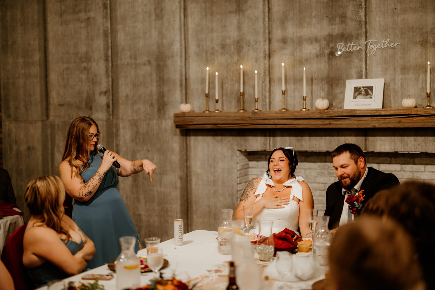 A woman in a teal dress speaks into a microphone as a laughing bride and smiling groom listen at a candlelit rustic wedding reception.