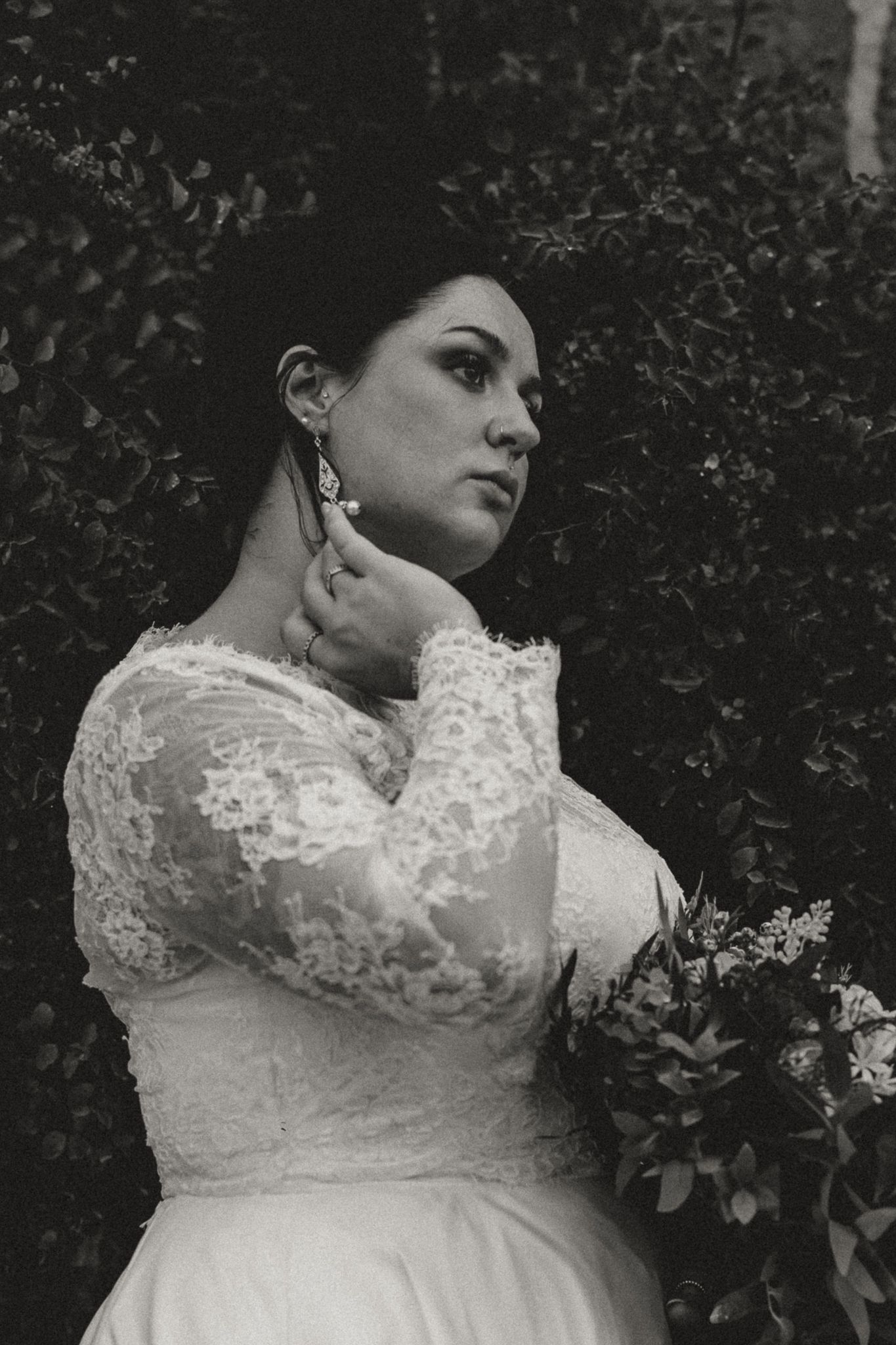 Black and white portrait of a bride in a lace long-sleeve wedding dress touching her earring while holding a bouquet near greenery.