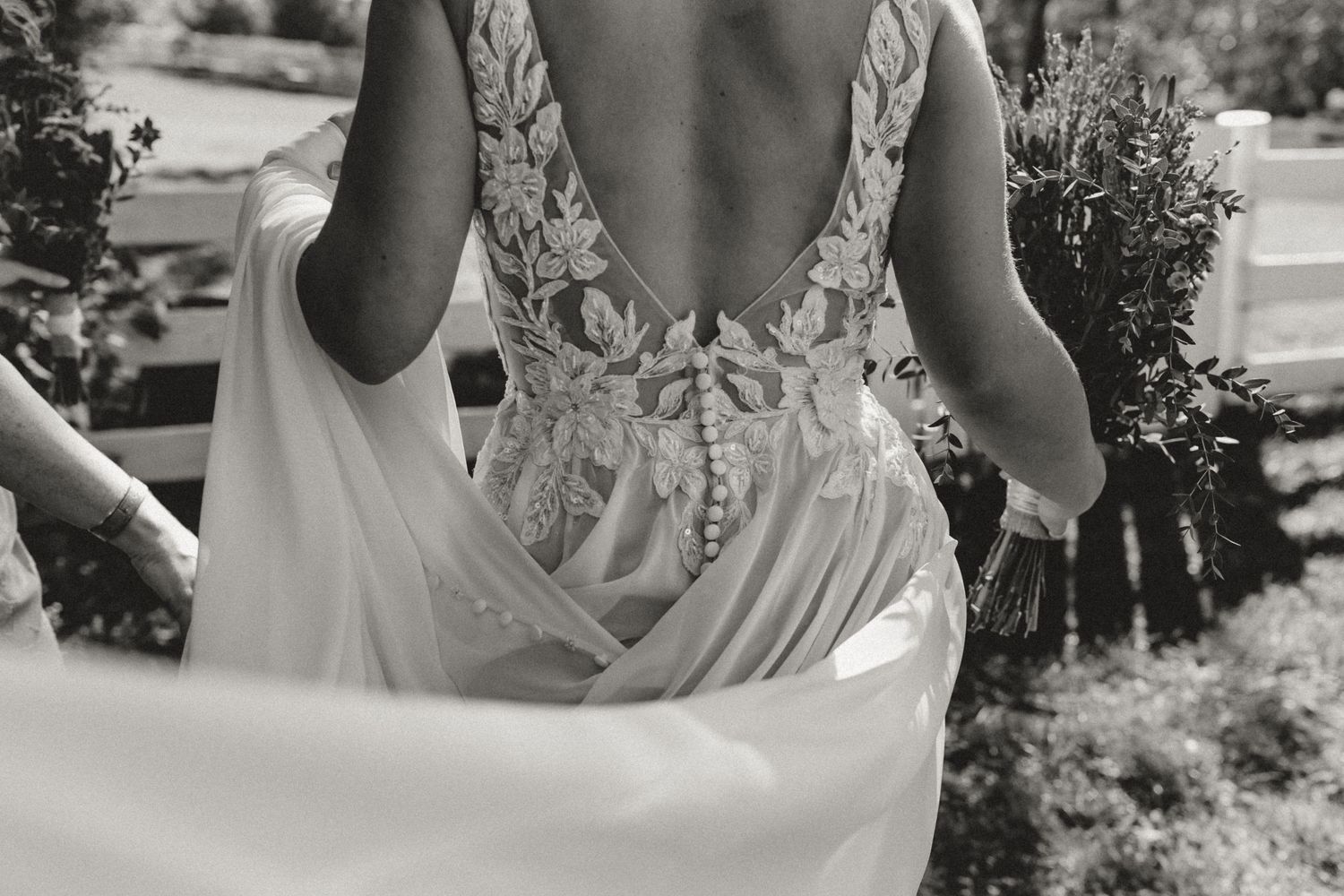 Black and white photo of a bride from behind in a lace open-back wedding gown with button detail, holding a bouquet outdoors.