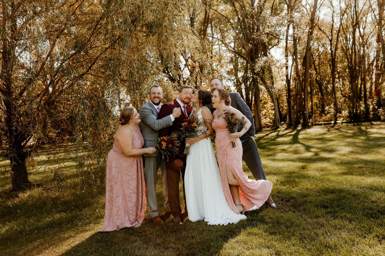 Bride, groom and four wedding party members in blush dresses and suits share a joyful group hug under willow trees in golden sunlight.