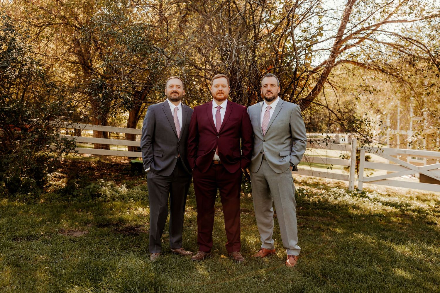 Three men in suits, one in burgundy between two in gray, stand together on a grassy lawn by a white wooden fence and autumn trees.