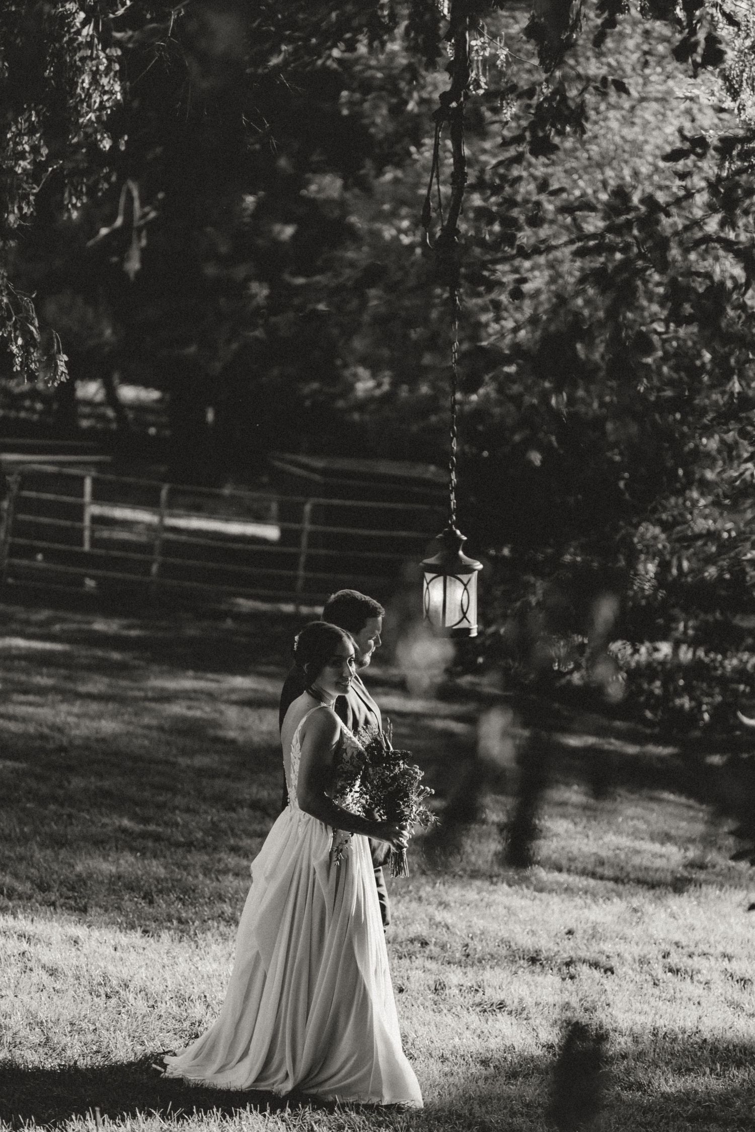 Black and white photo of a bride in a flowing gown holding a bouquet, standing beside a groom under a hanging lantern in a sunlit garden.