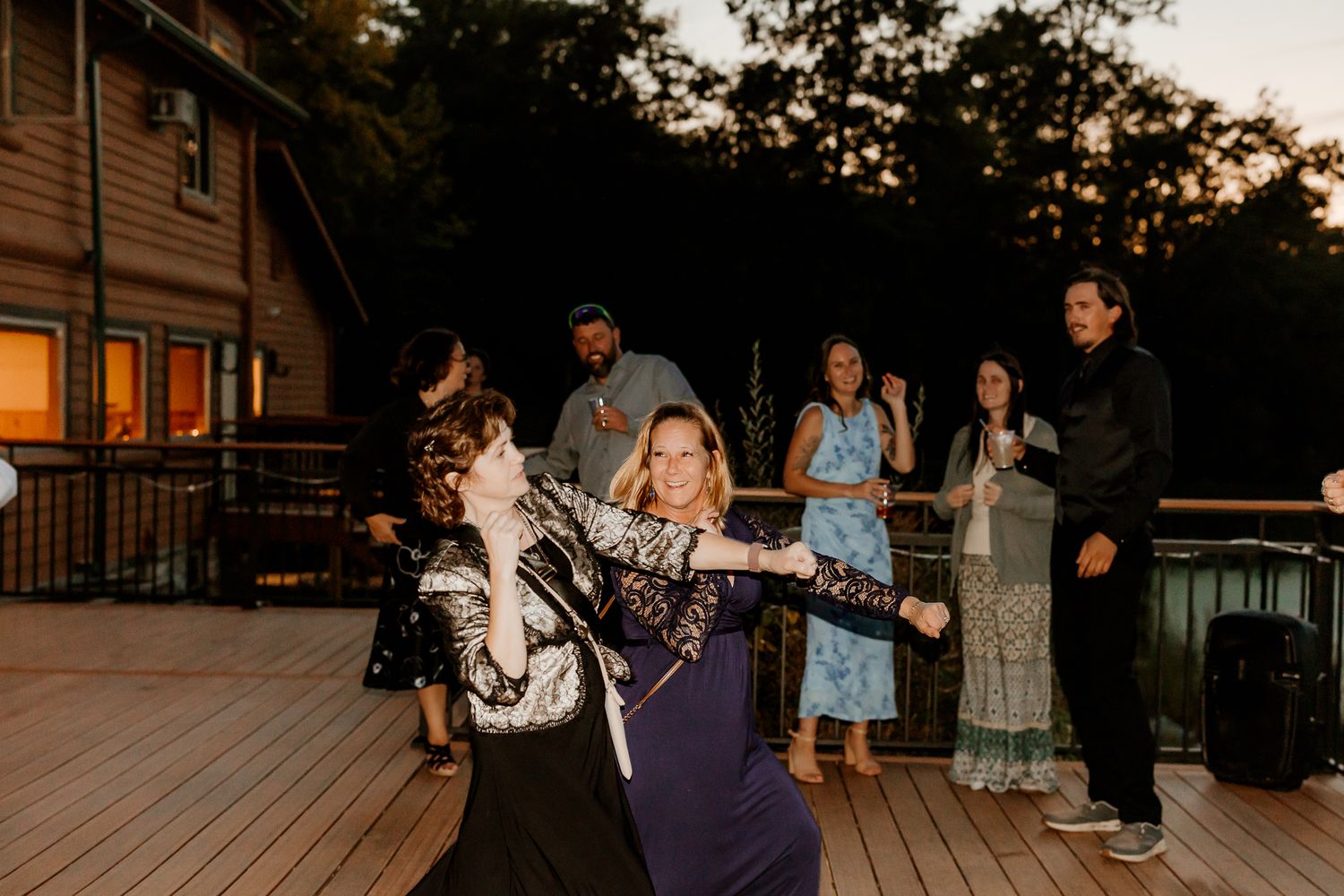 Wedding guests laugh and dance together on a wooden deck at dusk outside a rustic lodge, with trees and warm window light in the background.