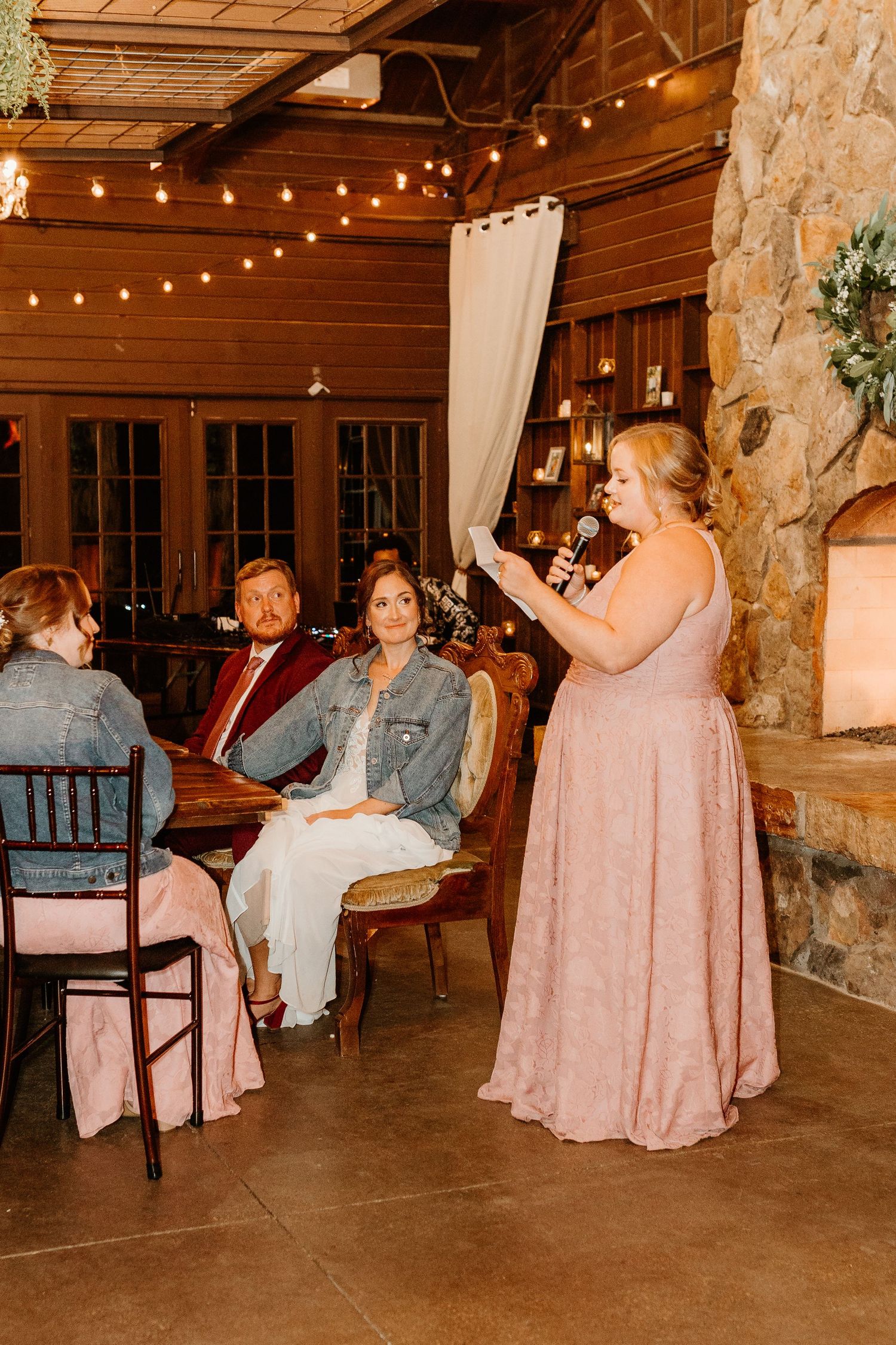 Bridesmaid in a blush pink lace gown gives a toast with a microphone to seated wedding guests in a rustic stone and wood venue.