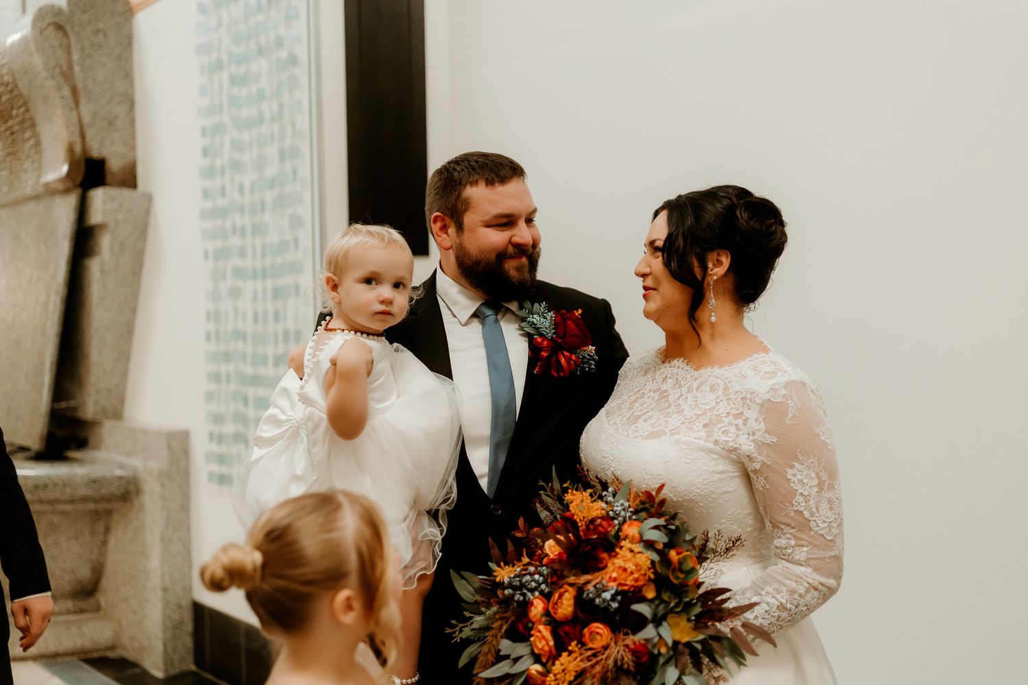 Bride in a lace gown with an autumn bouquet stands beside a smiling groom holding a baby girl in white, with a child in front.