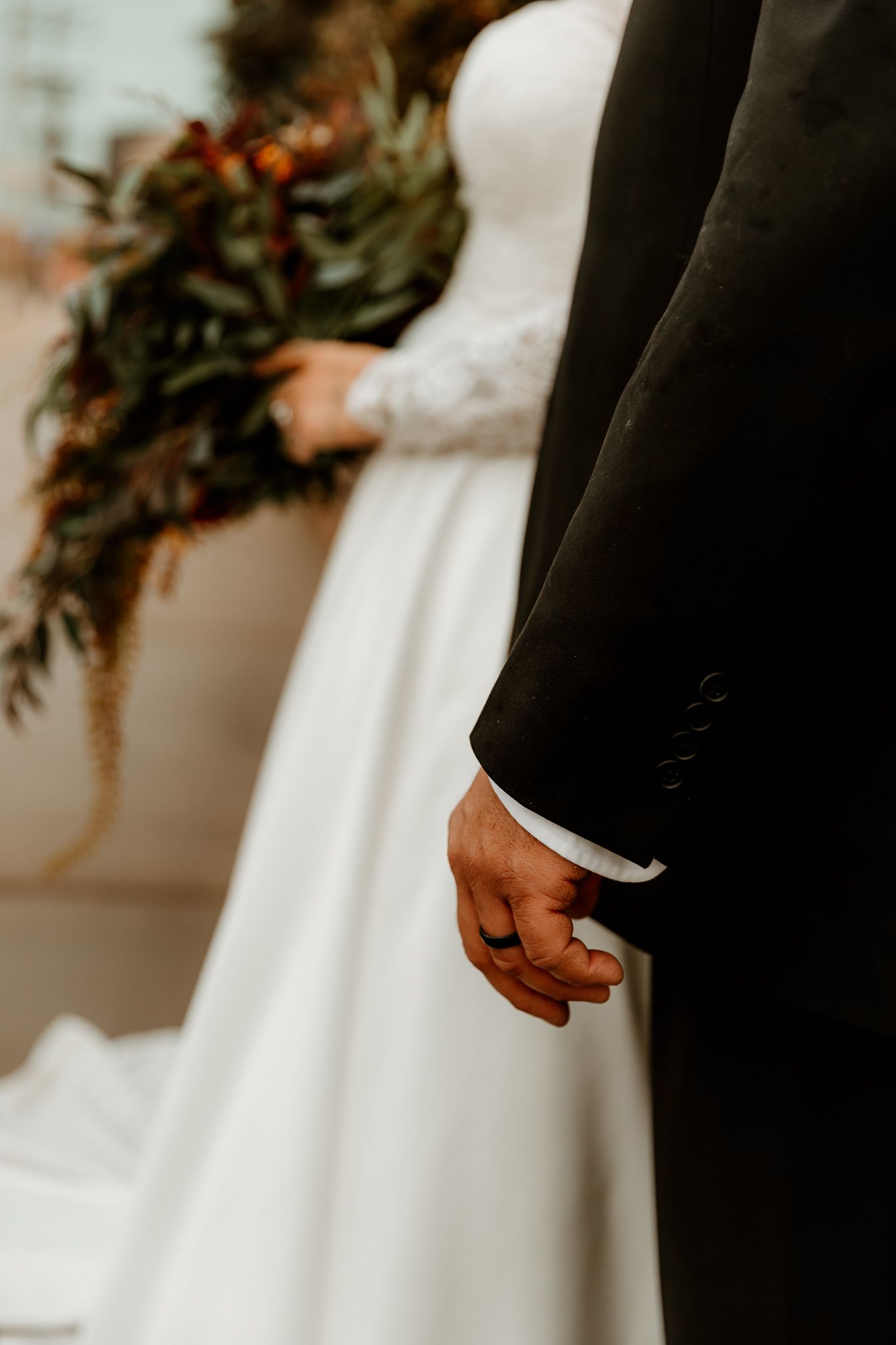 Groom in a black suit with a wedding ring stands beside a bride in a white lace-sleeved gown holding an autumn bouquet.