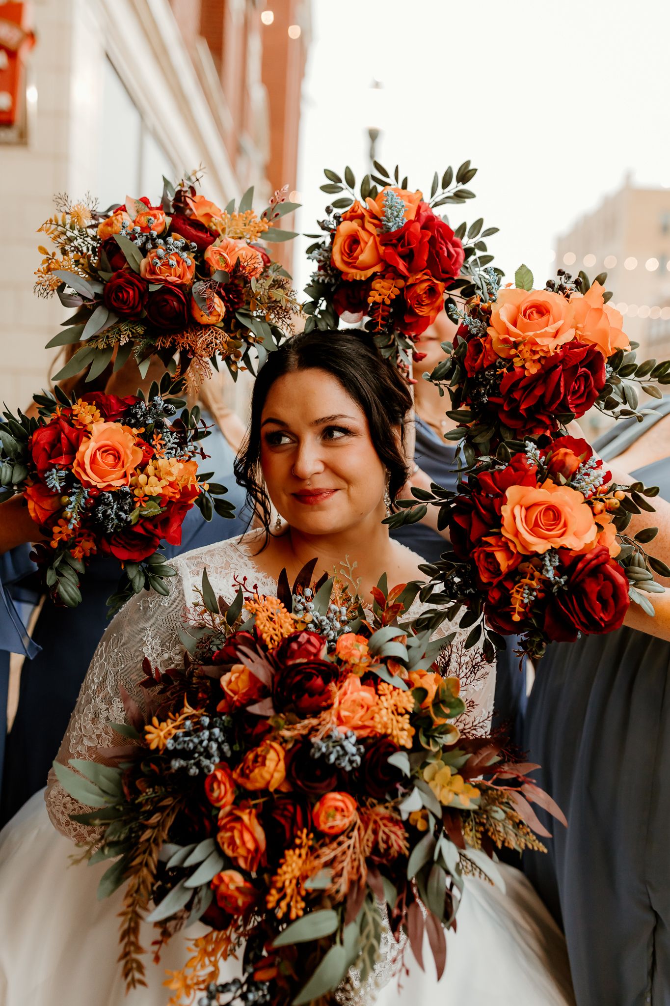 Smiling bride in a lace gown holds an autumn bouquet of orange and red roses, surrounded by bridesmaids' matching floral bouquets.