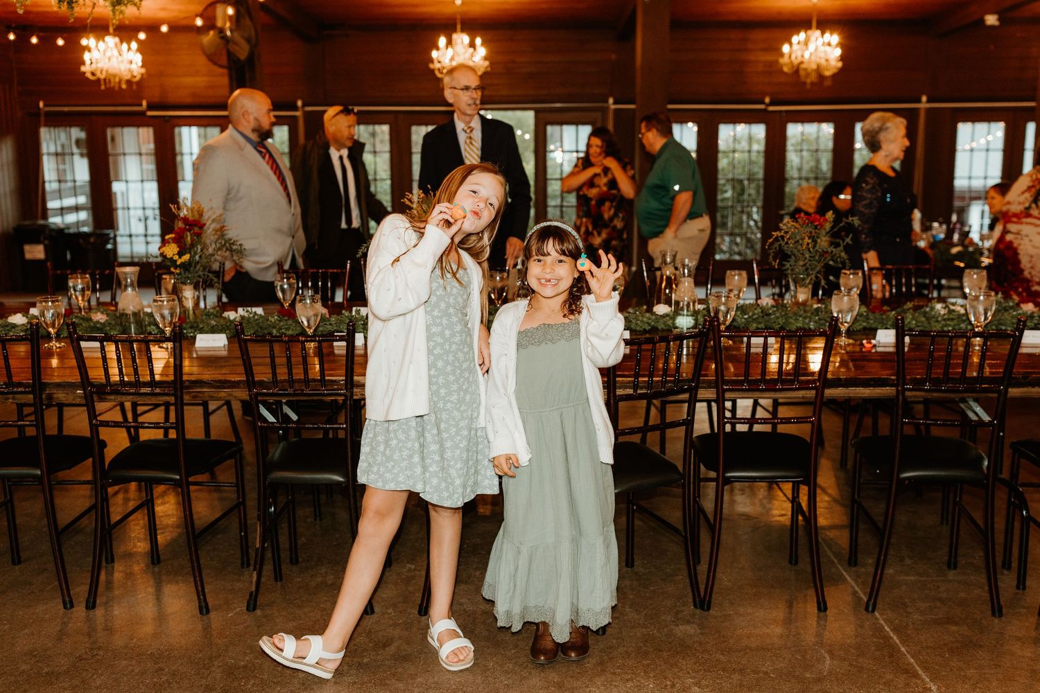 Two smiling girls in light dresses wave and pose in an elegant banquet hall with set tables and guests mingling in the background.
