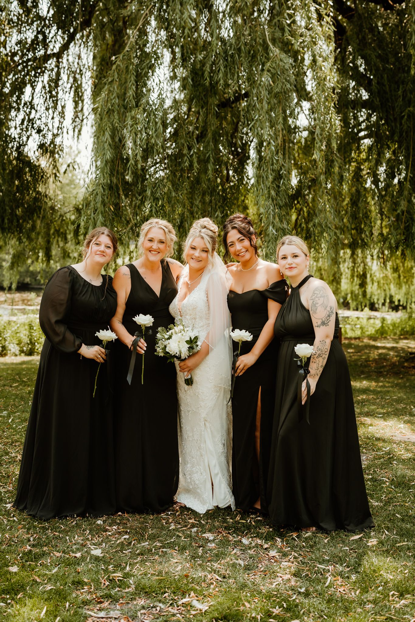 Bride in white gown poses with four bridesmaids in black dresses under a weeping willow tree.
