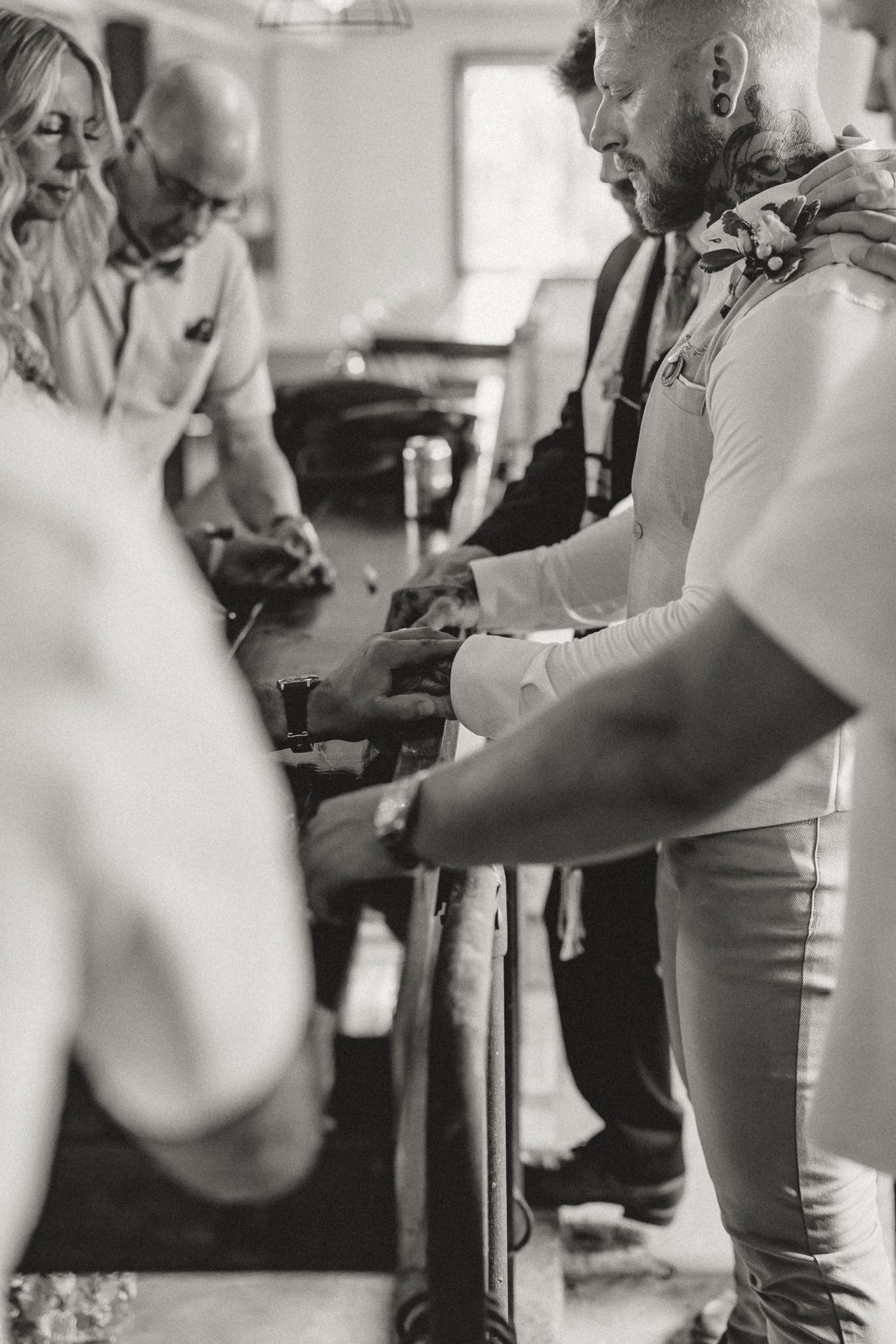 Black and white photo of groomsmen preparing for a wedding ceremony, adjusting accessories at a table.