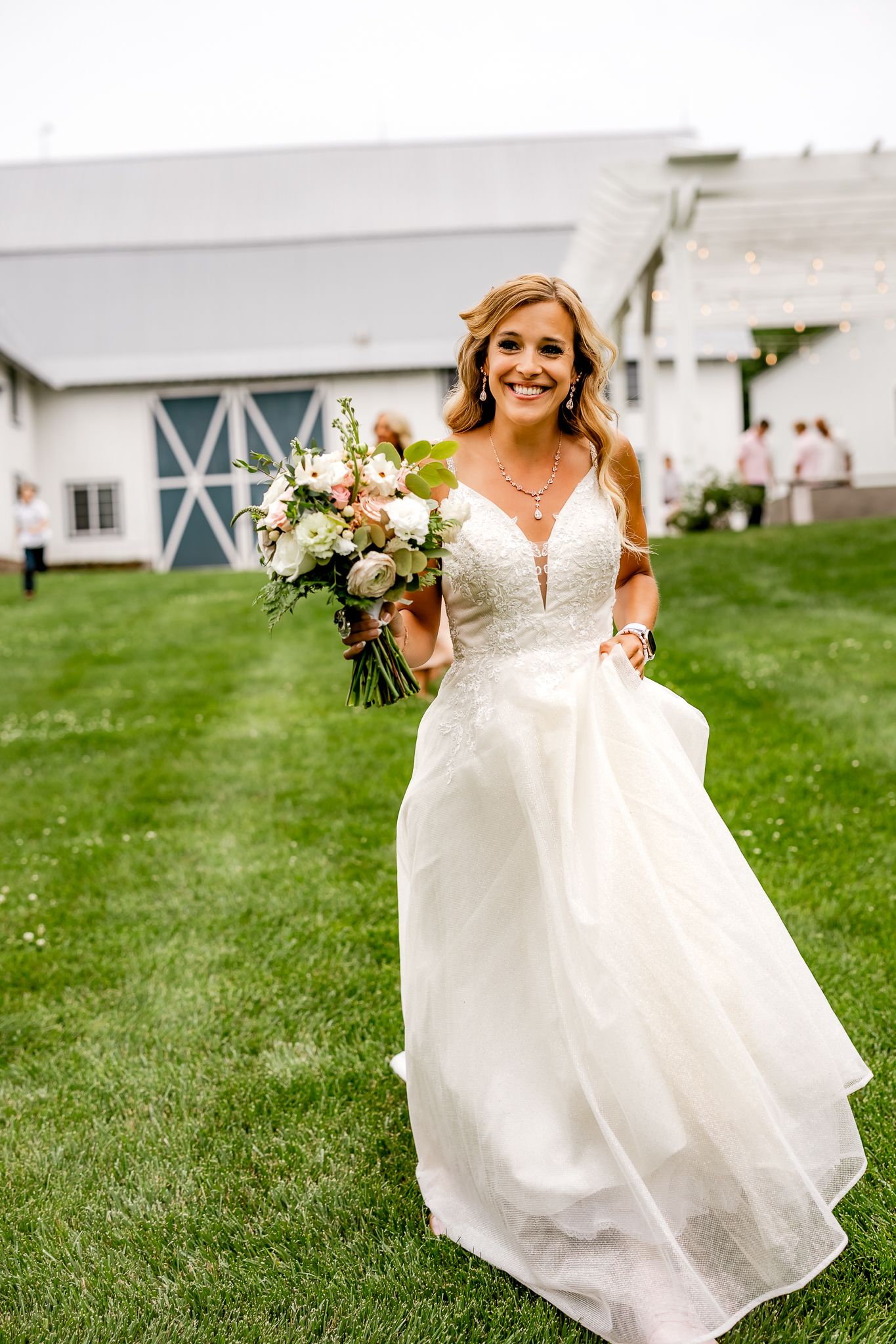 Smiling bride in white lace ball gown holding floral bouquet walks across green lawn near white barn venue.