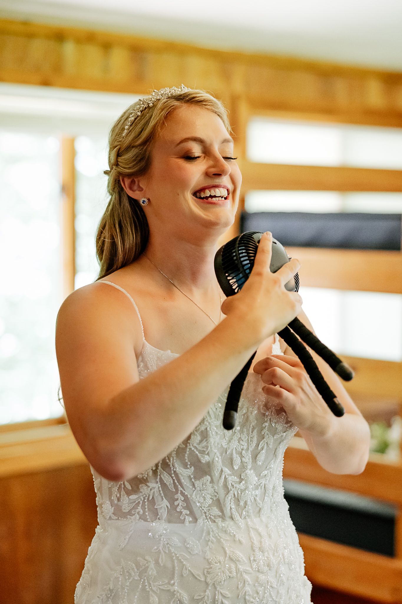 Laughing bride in white lace gown and tiara holds black heels while getting ready on her wedding day.