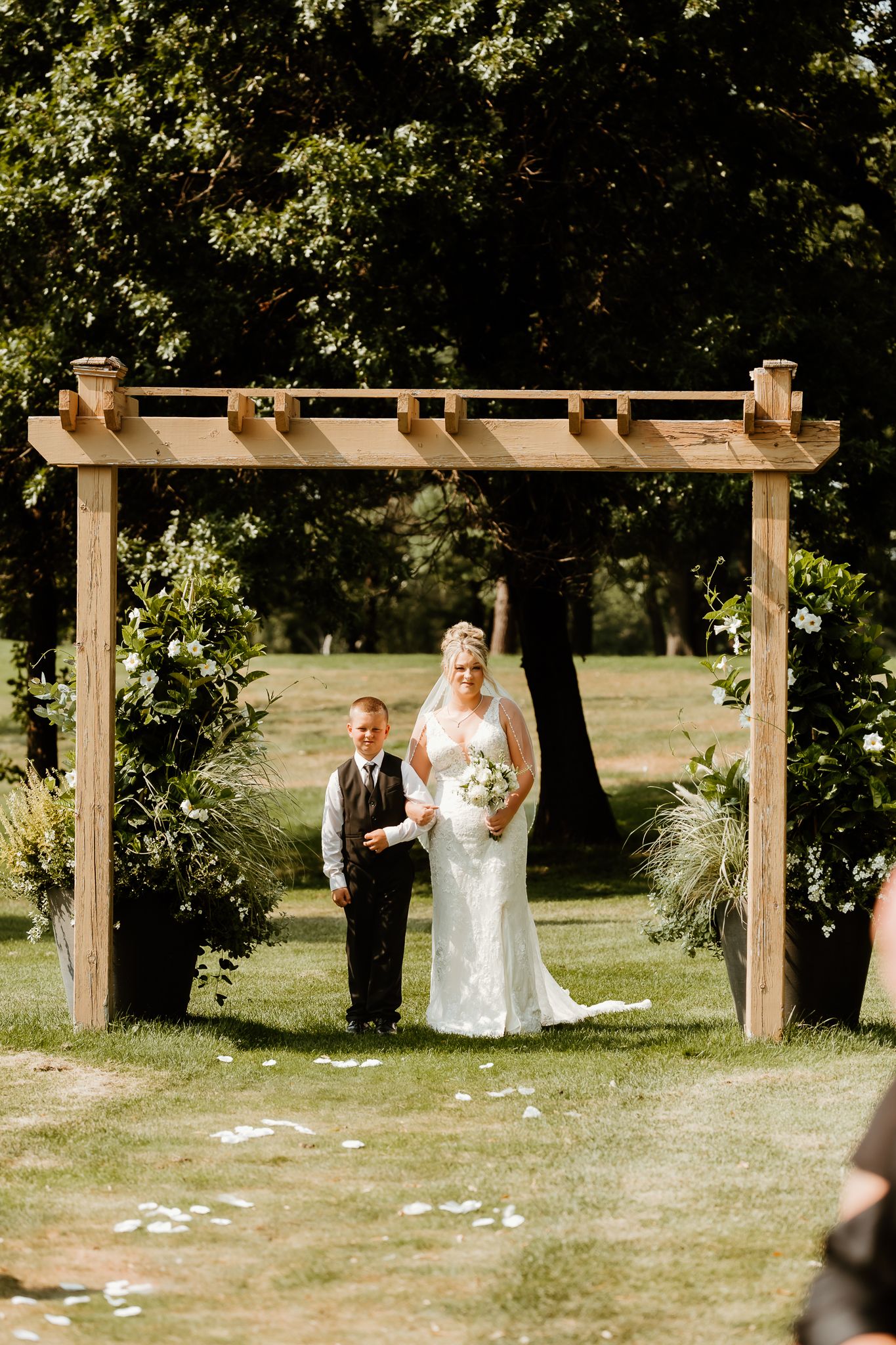 Bride in white lace gown holding bouquet stands with young boy in suit under wooden pergola at outdoor wedding ceremony.