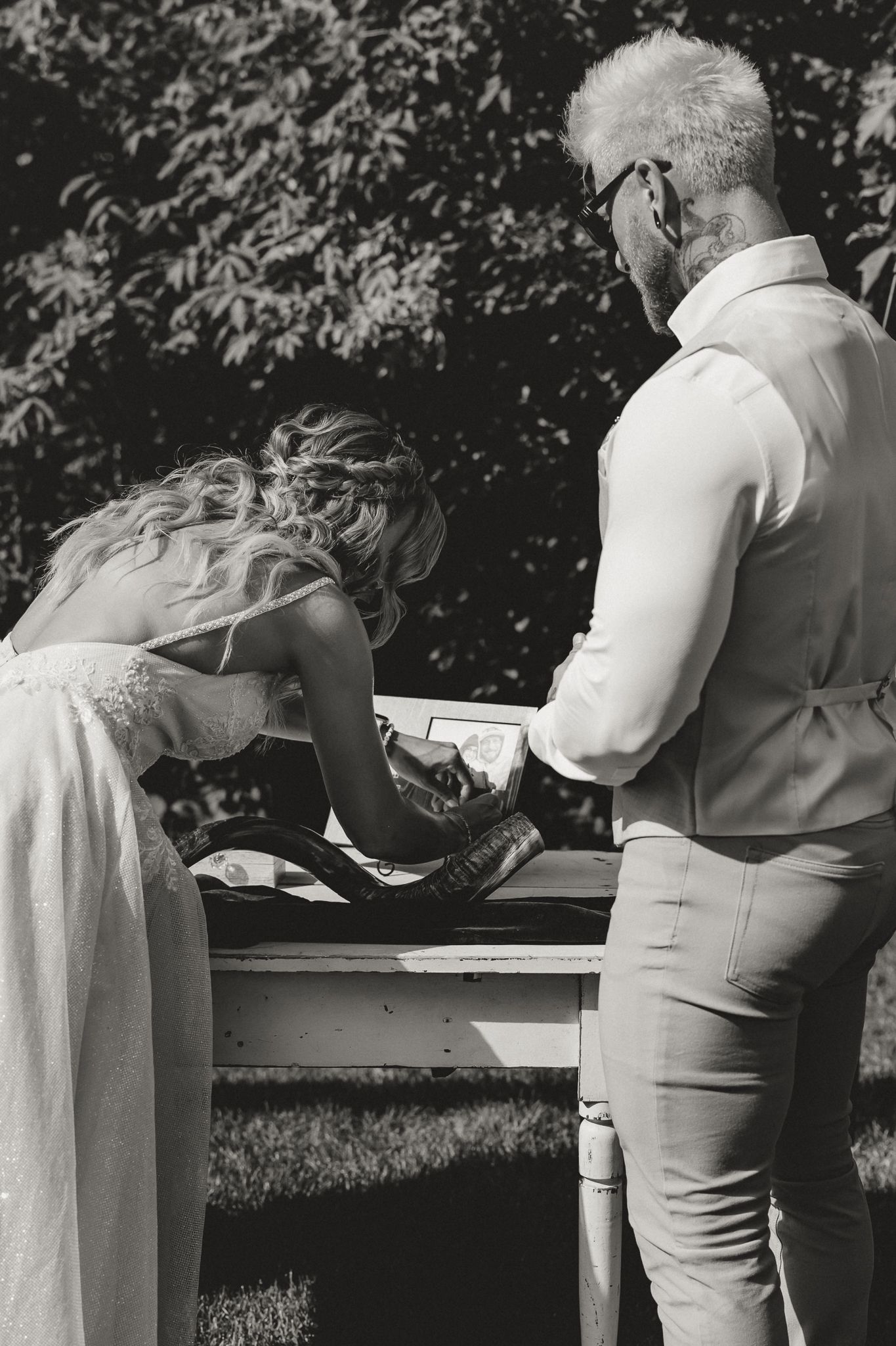 Black and white photo of bride and groom signing wedding documents outdoors near lush green trees.