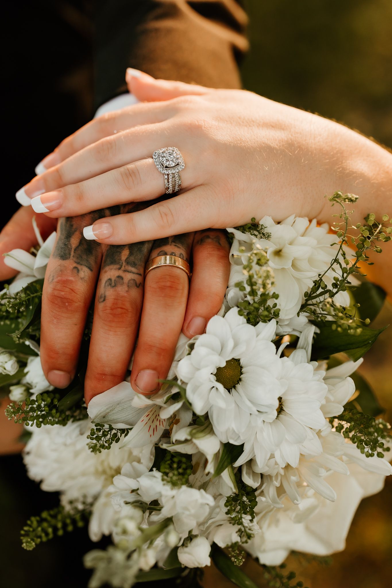 Newlyweds display wedding rings on tattooed and manicured hands resting on a white floral bouquet.
