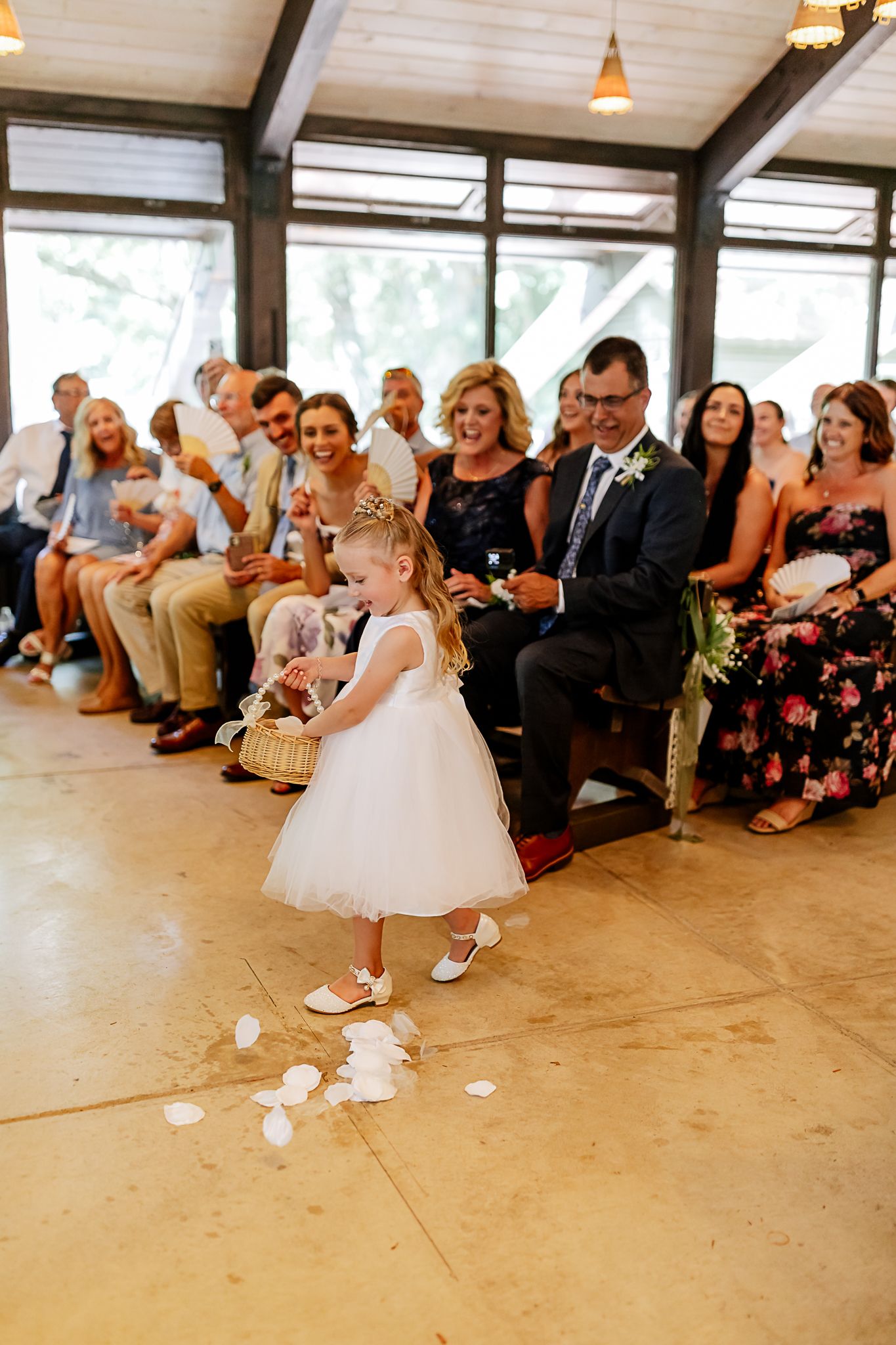 Flower girl in white dress scattering rose petals down the aisle at a wedding ceremony.