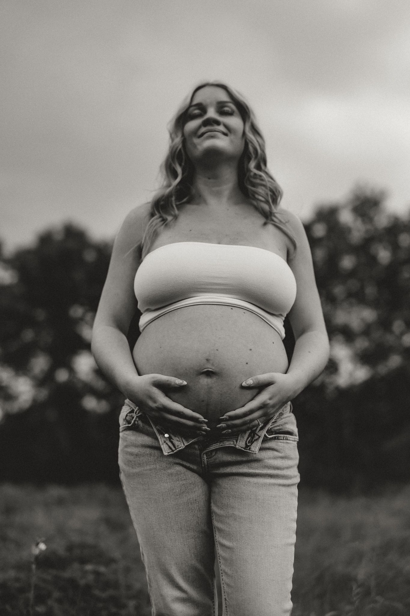 Black and white maternity portrait of joyful pregnant woman holding belly in outdoor field setting.
