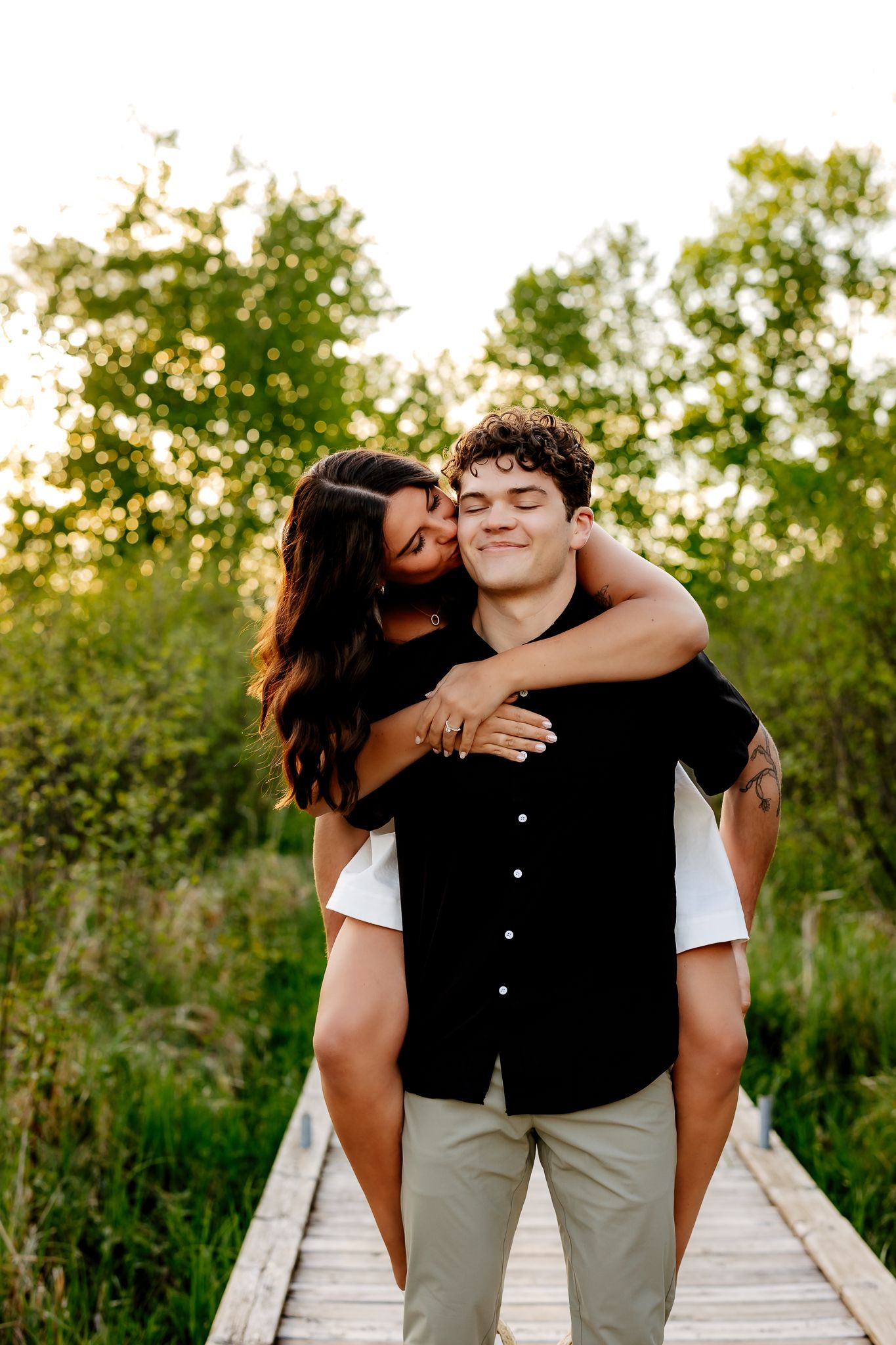 A young couple shares a romantic piggyback ride on a wooden boardwalk surrounded by lush green trees at golden hour.