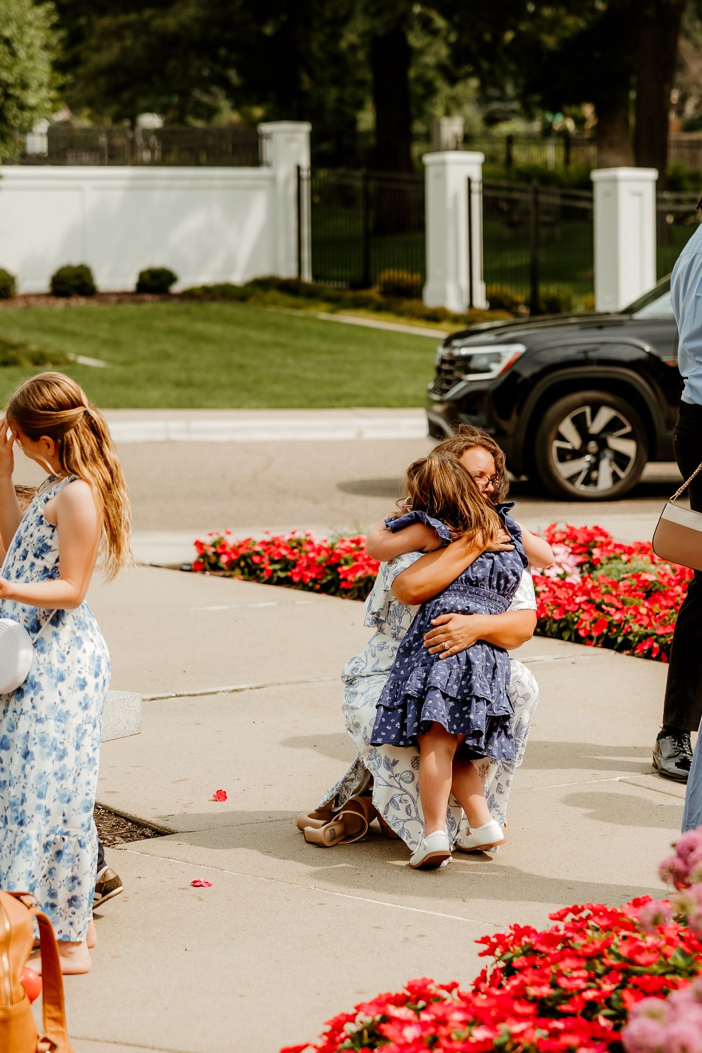 Two young girls in blue dresses share a warm hug outdoors near red flower beds at a garden event.