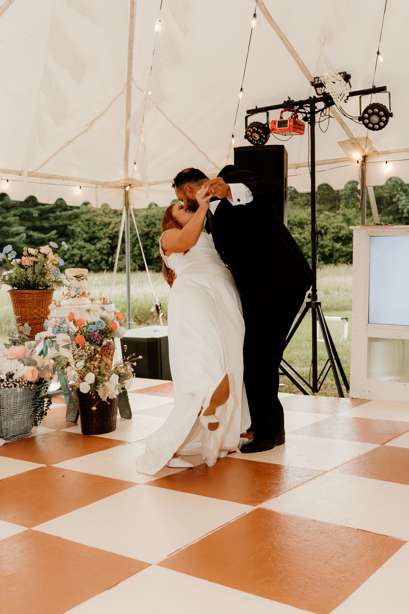 Bride and groom share first dance kiss on checkered dance floor under tent with fairy lights and floral arrangements.