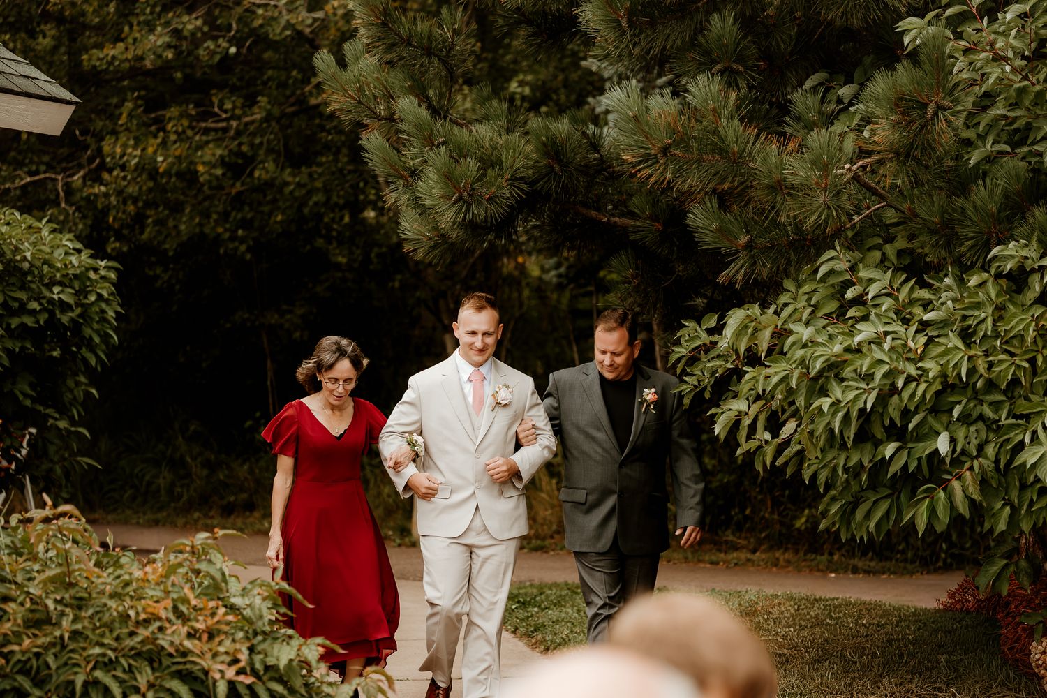 Groom in light suit walks with parents through garden path at outdoor wedding ceremony.