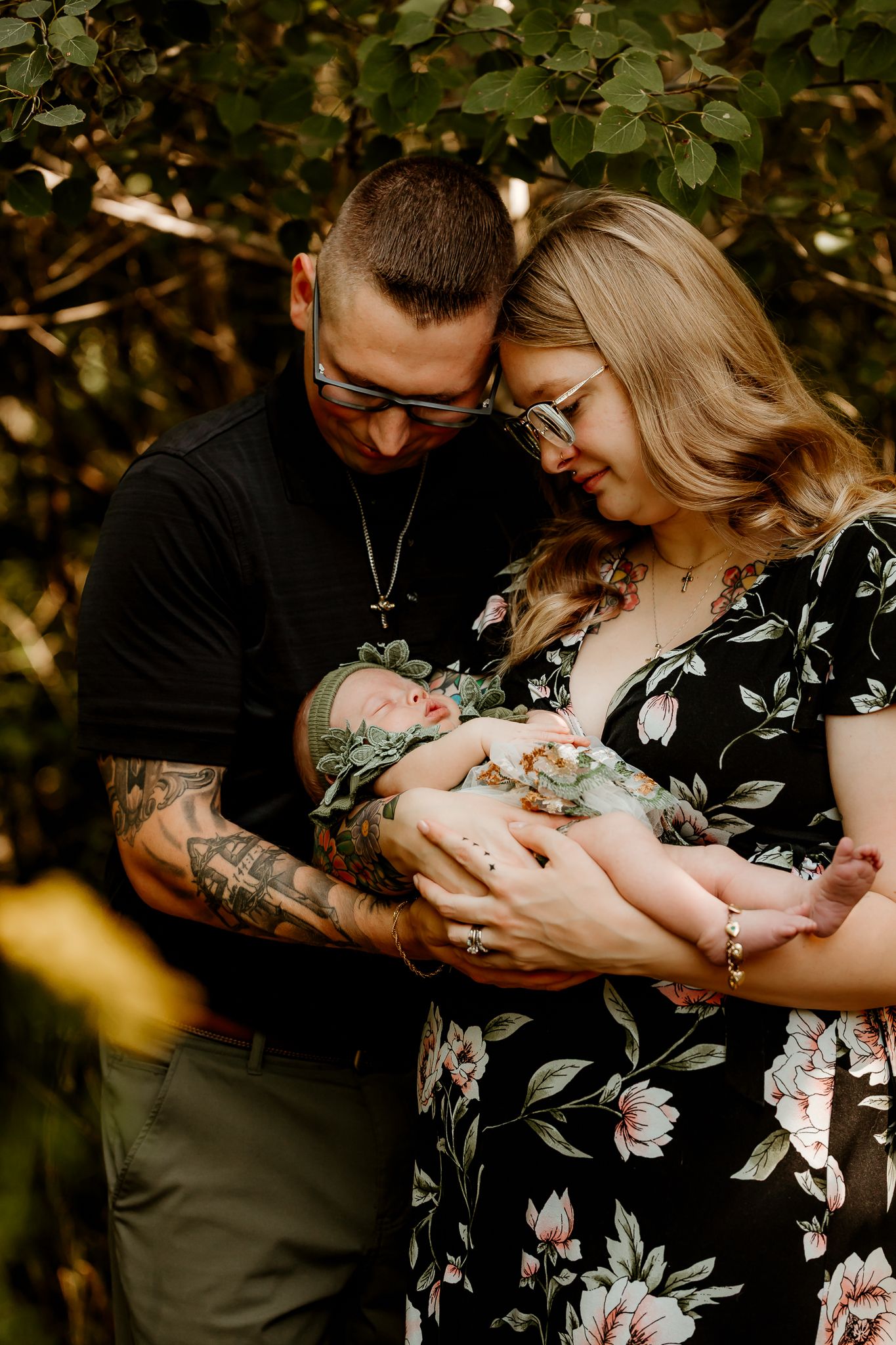 A couple tenderly holds their newborn baby outdoors, surrounded by lush green foliage in a heartfelt family portrait.