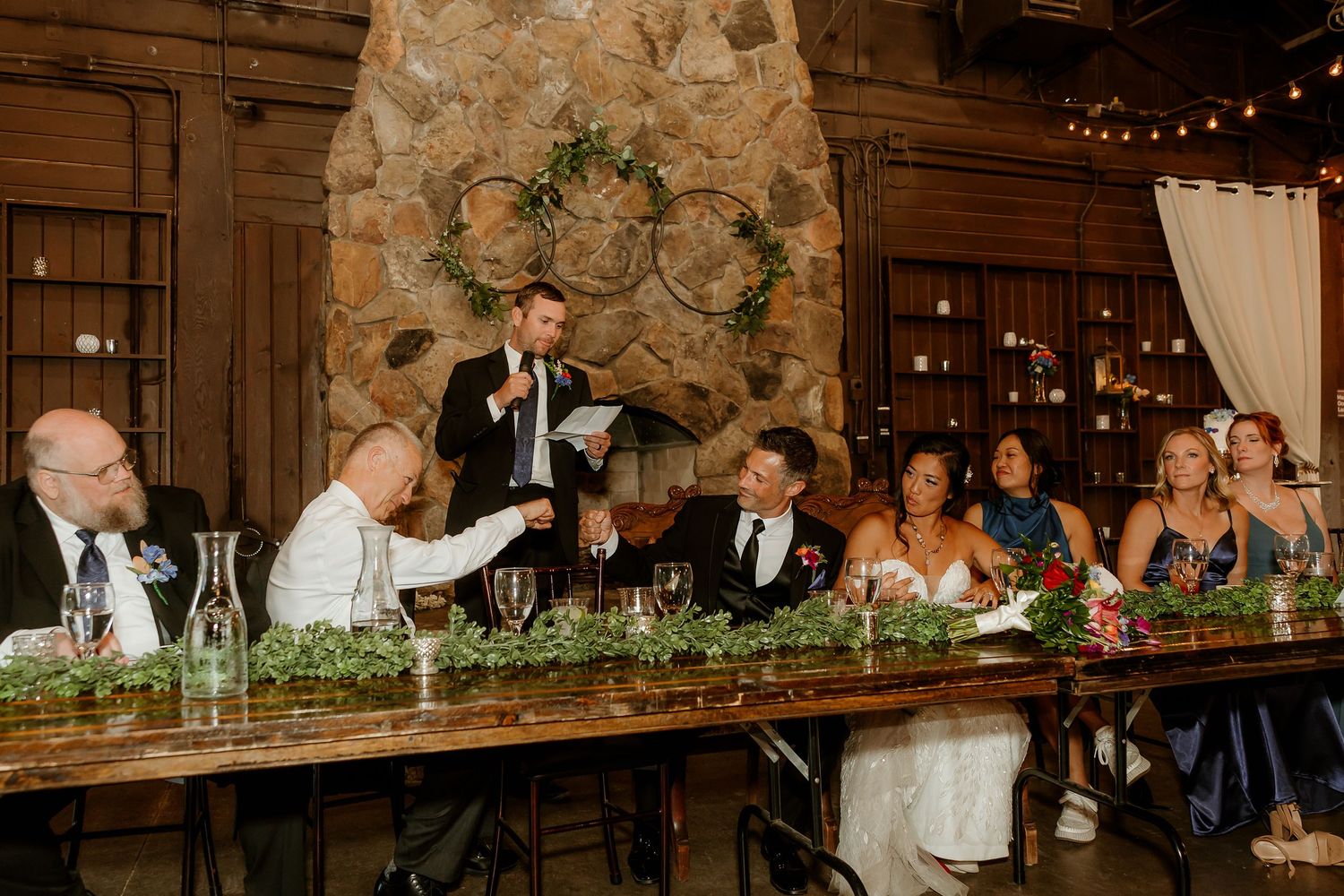 A groomsman delivers a heartfelt wedding toast at a rustic stone venue while the bride and groom sit at a decorated head table.