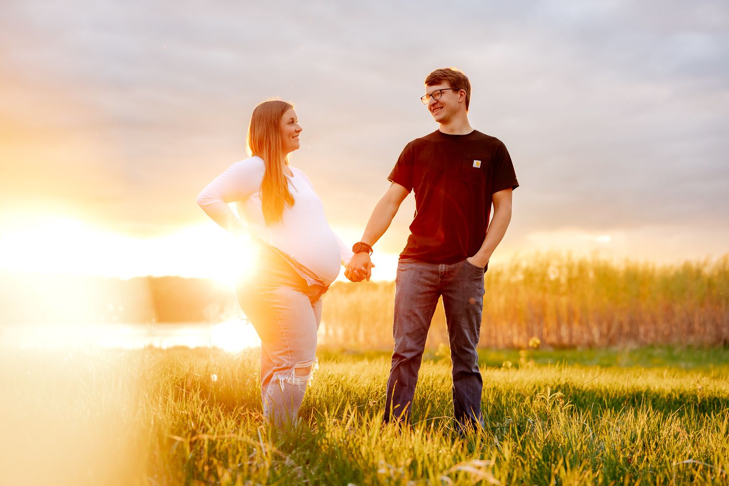 A pregnant woman and her partner hold hands in a golden sunlit field at sunset, smiling at each other.