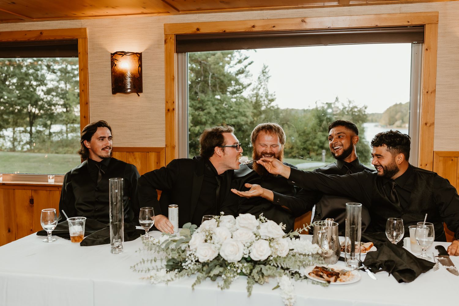 Five men in black shirts laugh and reach toward each other at a wedding reception table with white floral centerpieces and a lakeside view.