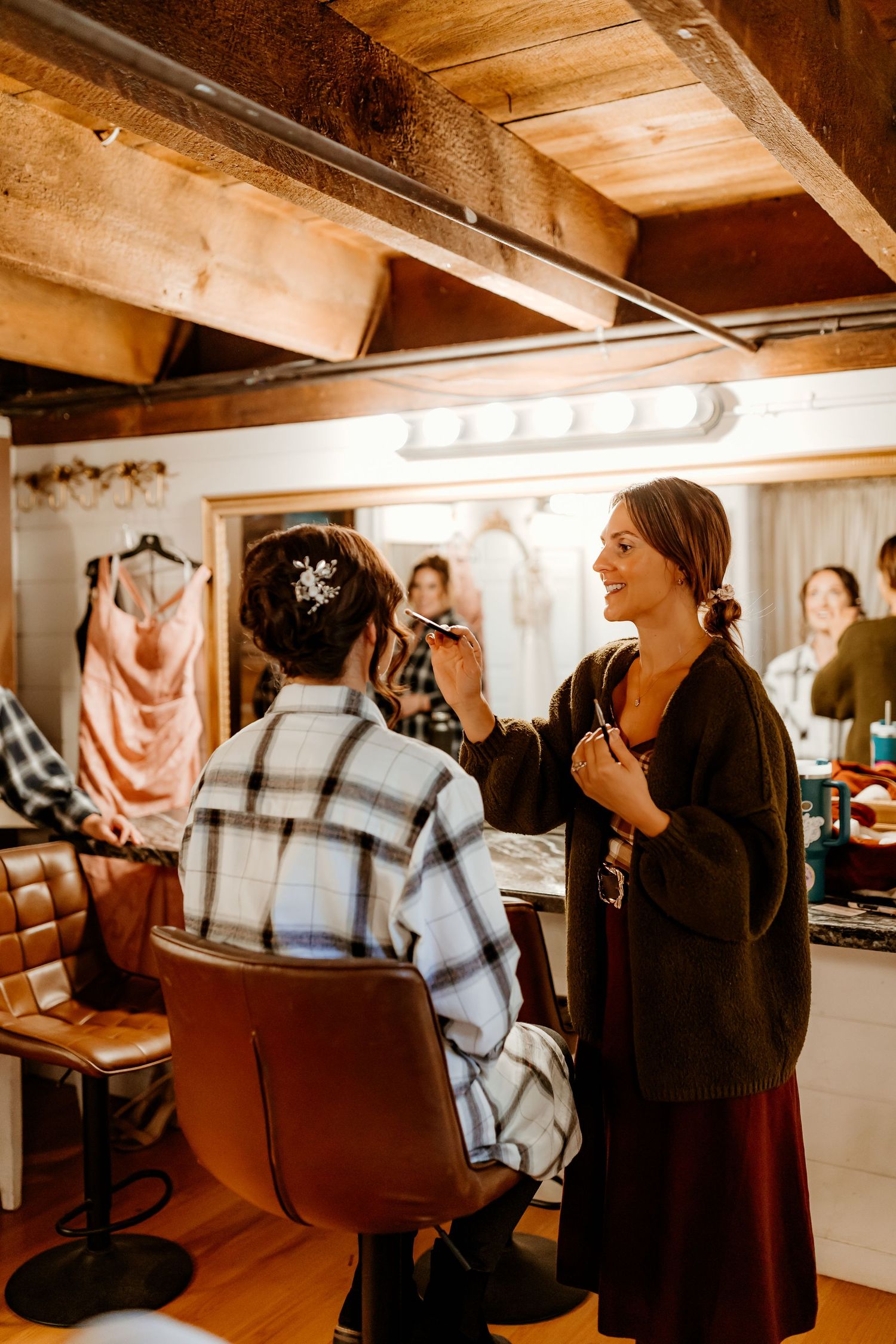 A makeup artist in a dark cardigan applies makeup to a seated woman in a plaid shirt with a floral hair clip in a rustic salon.