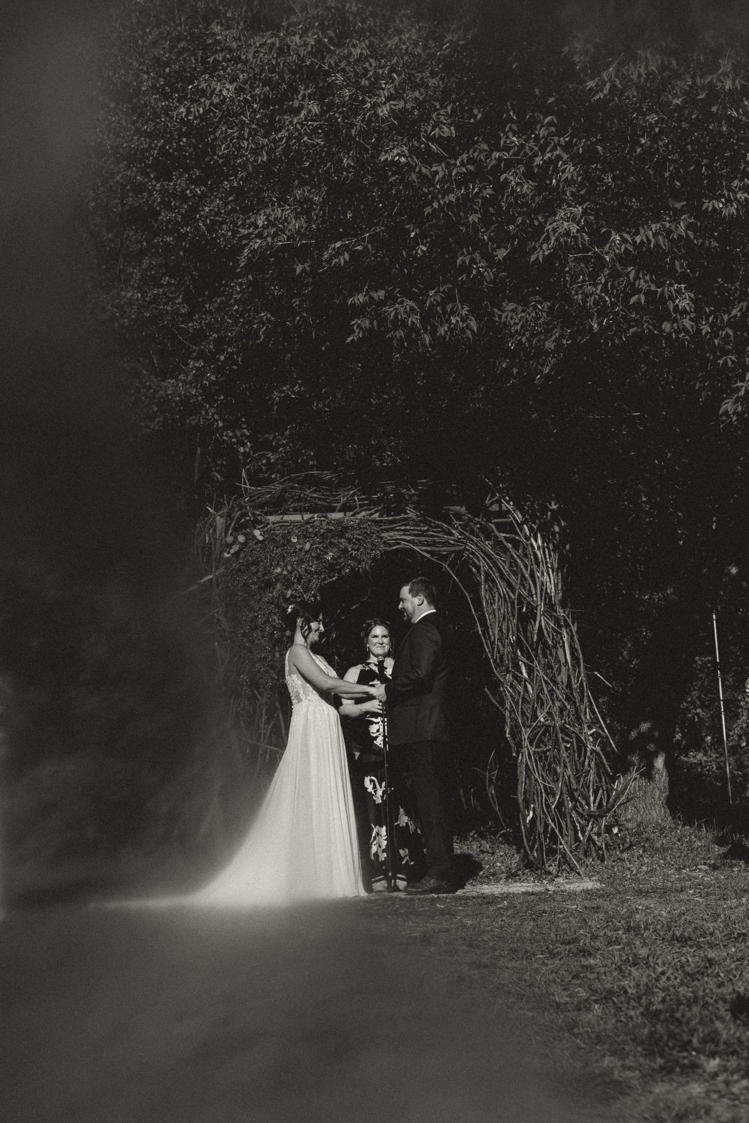 Black and white photo of a couple holding hands during an outdoor wedding ceremony beneath a rustic woven branch arch with an officiant.