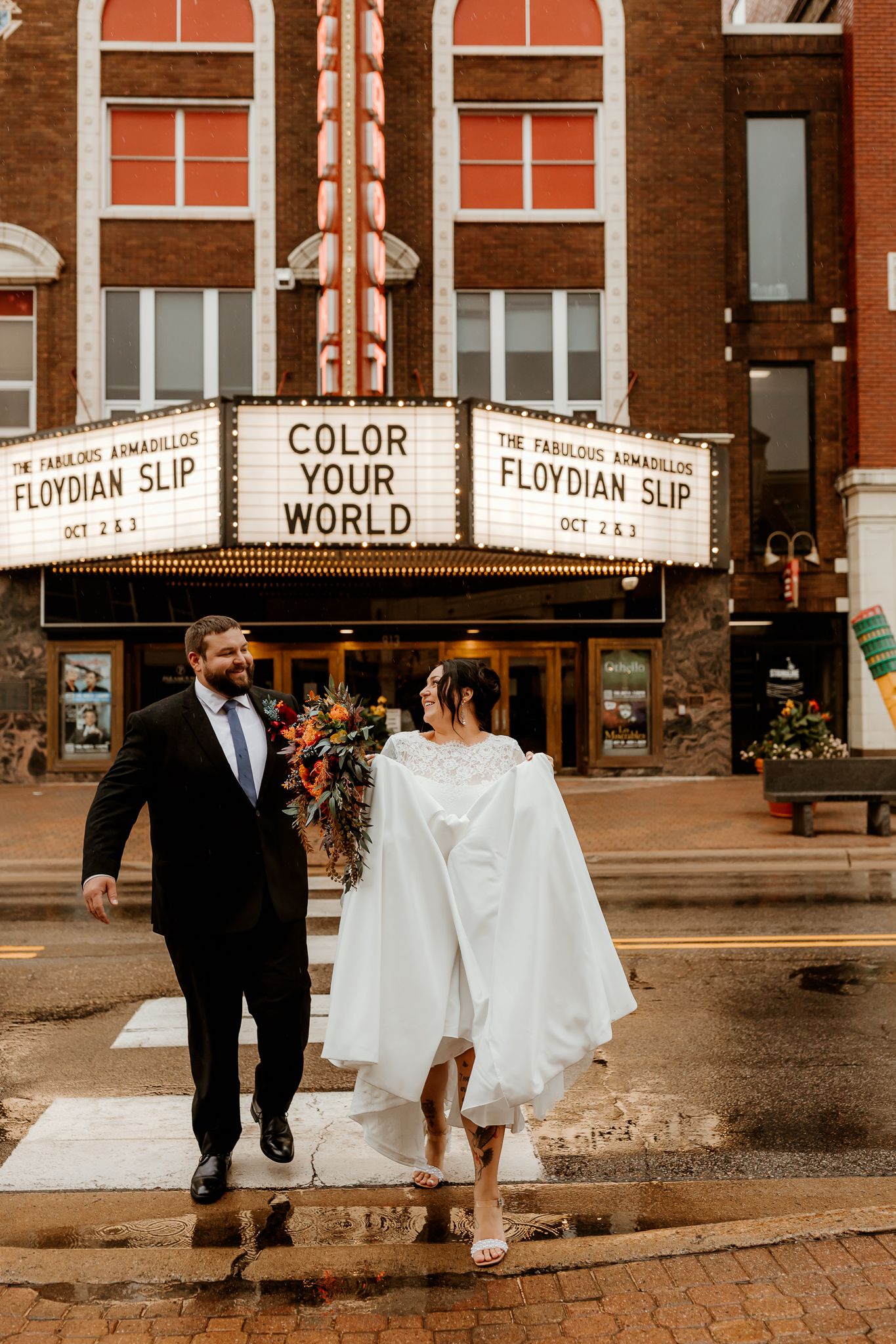 Bride in a white gown and groom in a dark suit with a bouquet cross a wet street in front of a vintage theater marquee reading Color Your Wo
