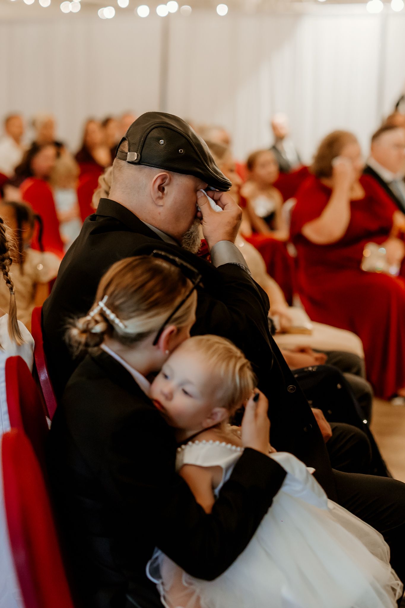 Emotional man in a cap wipes his tears while a woman holds a sleepy toddler in a white tulle dress during a wedding ceremony with guests.