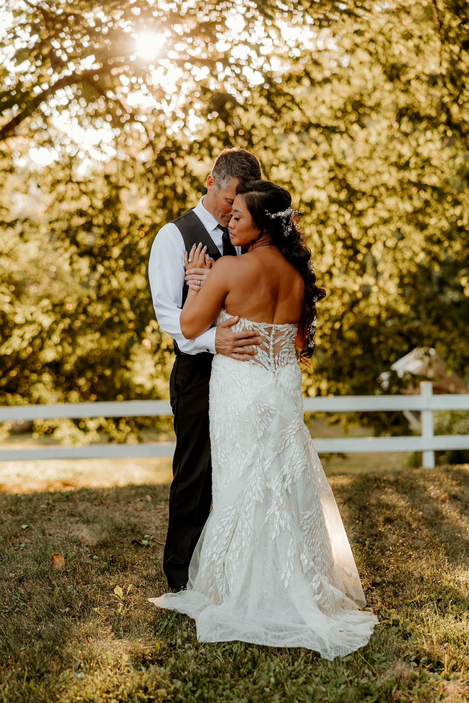 A bride and groom share a romantic embrace outdoors at golden hour, surrounded by lush trees near a white fence.