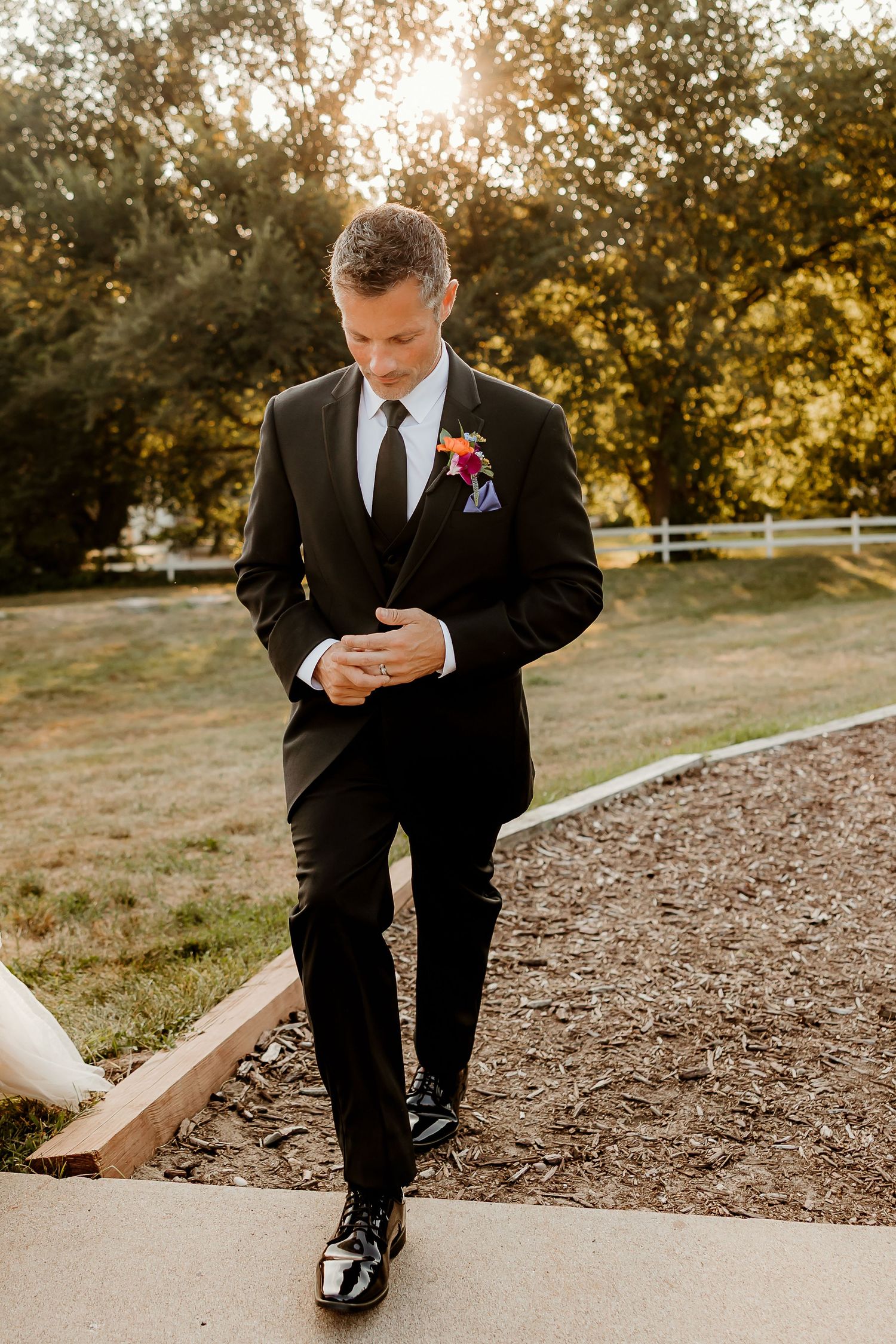 Groom in black tuxedo with colorful boutonniere adjusting cufflinks on garden path at golden hour.