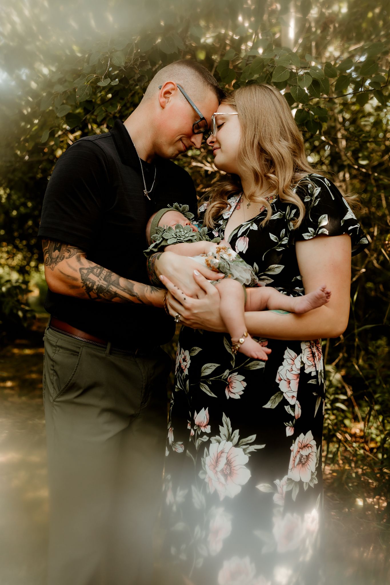 A couple with tattoos tenderly holds their newborn baby outdoors, touching foreheads in a loving embrace.