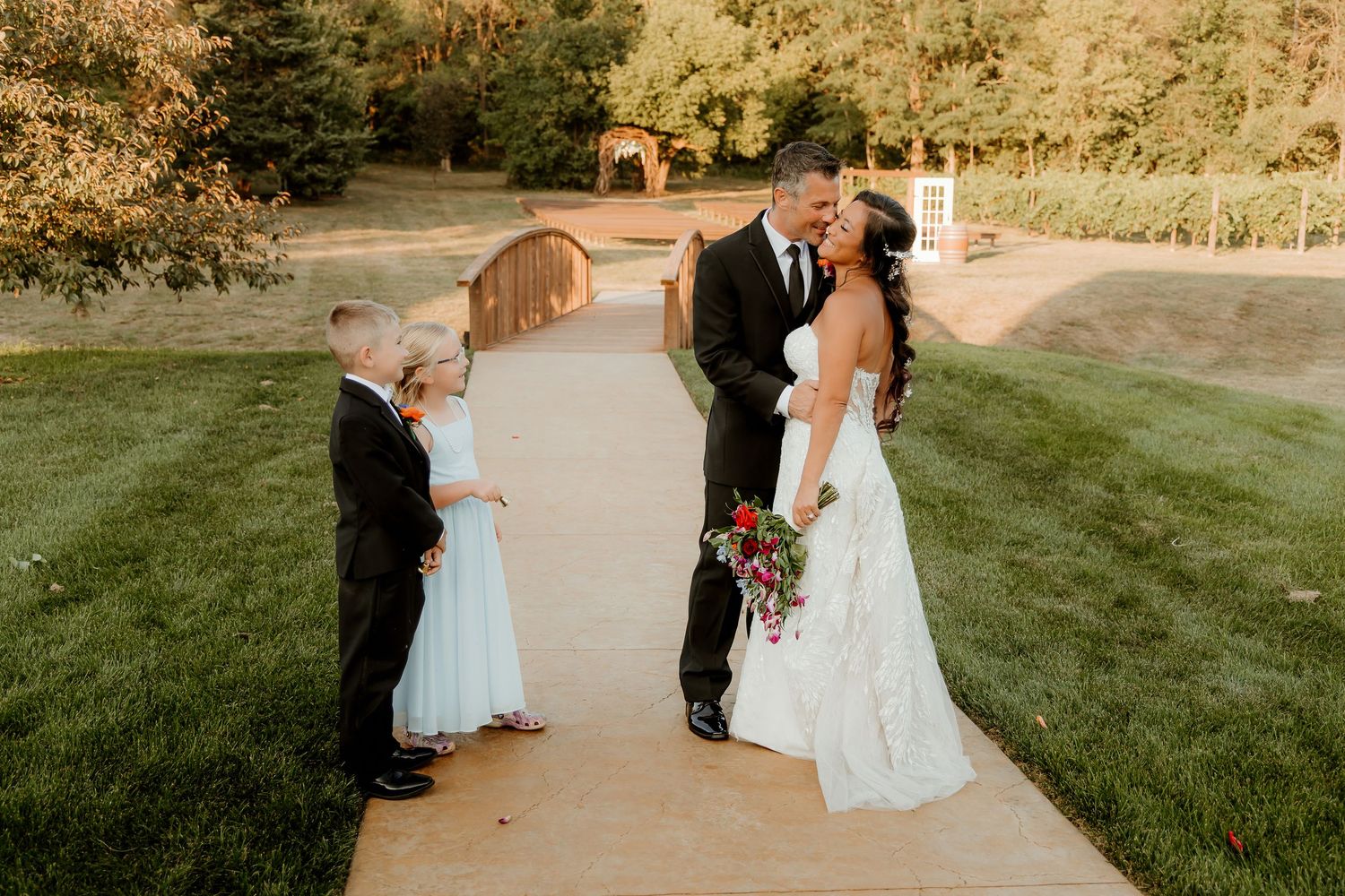 Bride and groom share a romantic moment on a garden path with flower girl and ring bearer at an outdoor wedding.