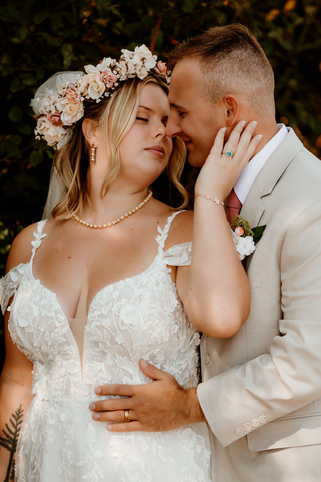 Bride in lace gown and floral crown shares intimate moment with groom in tan suit during outdoor wedding.