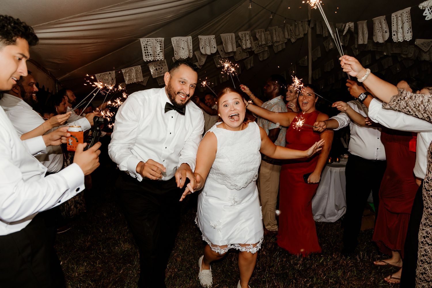 Bride and groom run through sparkler exit surrounded by cheering guests at outdoor wedding reception.