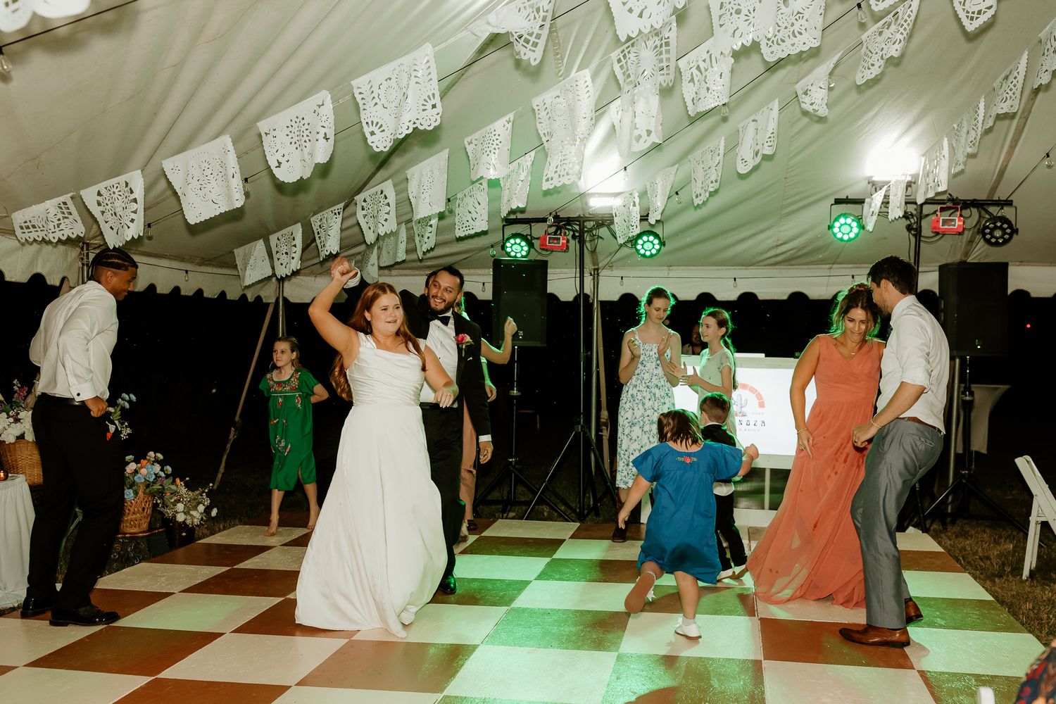 Bride and groom dancing with guests at a wedding reception under a white tent with decorative papel picado banners.