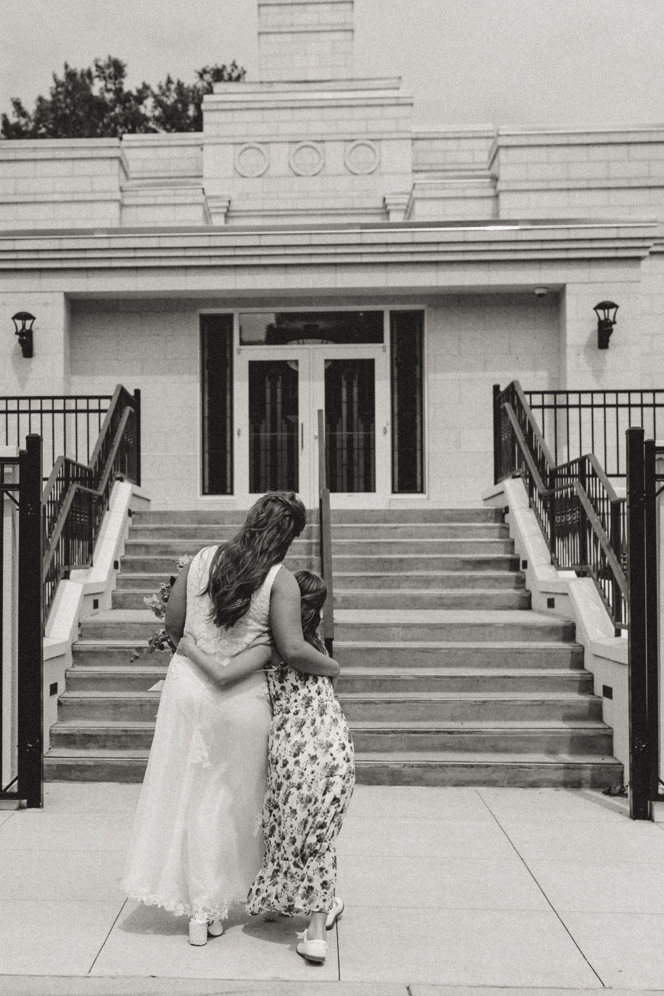 Two women in dresses walk arm in arm toward a white building with stairs in a black and white photo.